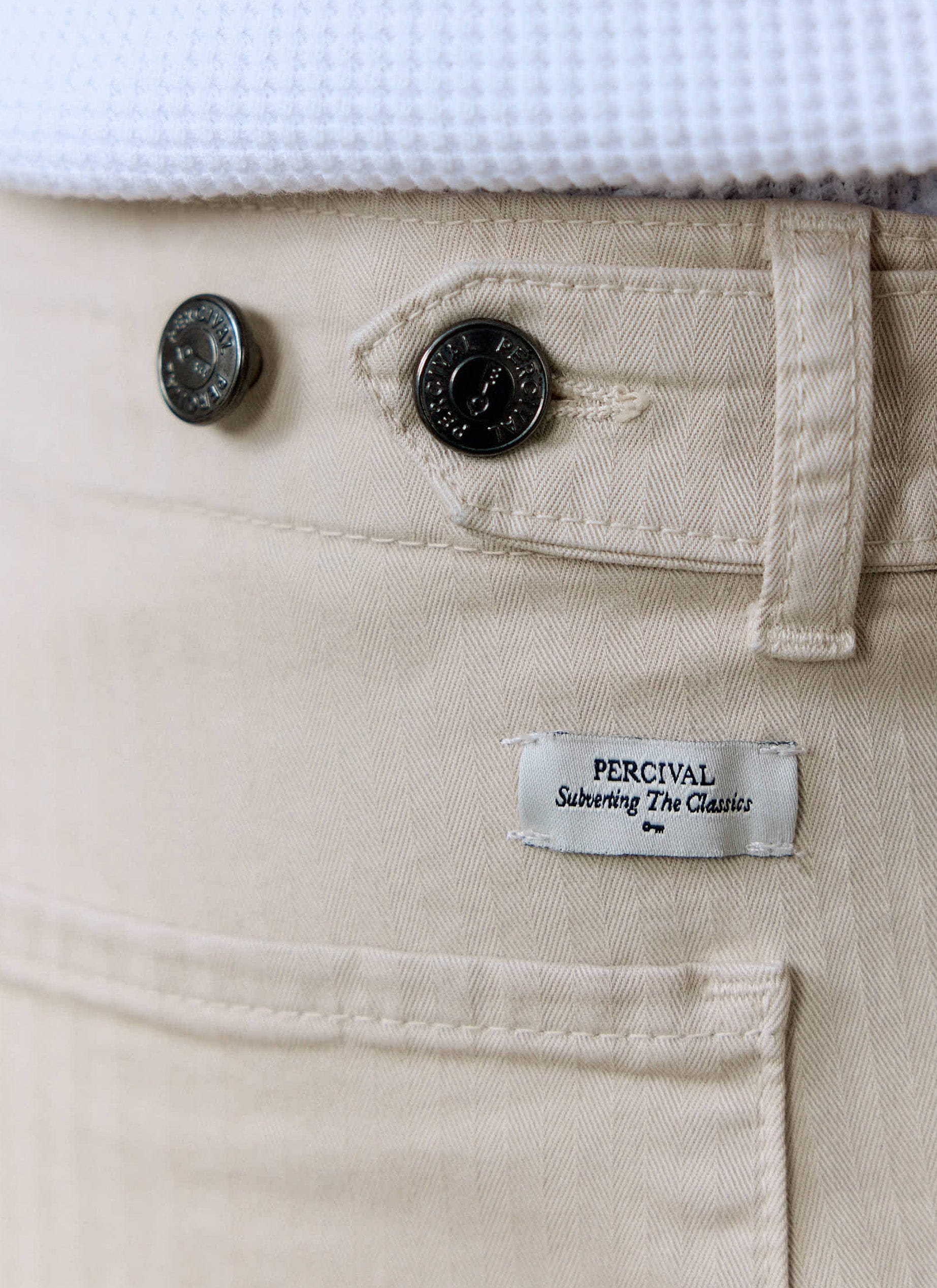 Close-up of the Percival Lungo Herringbone Straight Trousers in ecru, showing the branded buttons and logo label.