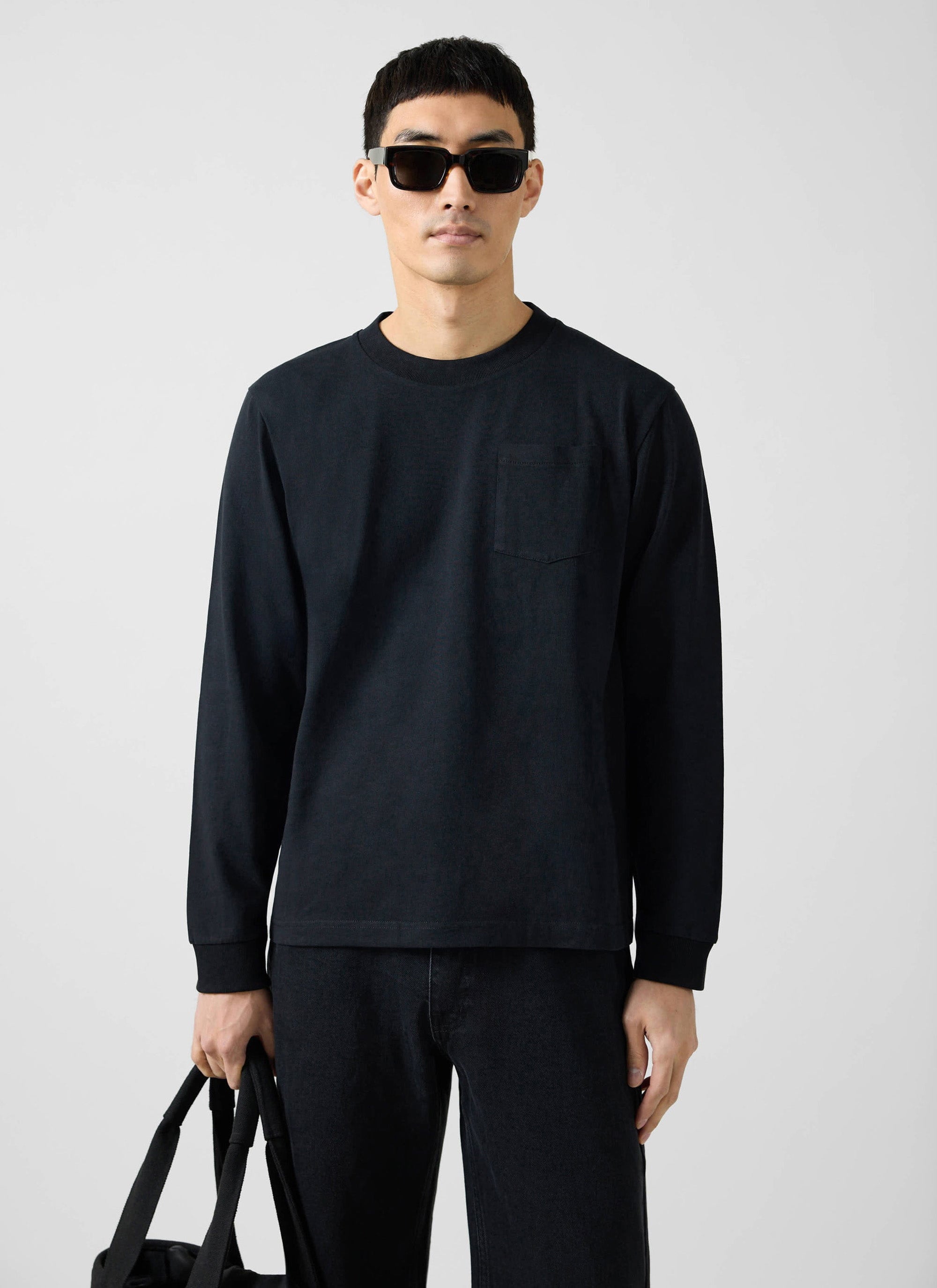 A model wears the Percival Long Sleeve Pocket T-shirt in black with matching black trousers, sunglasses and a tote bag.