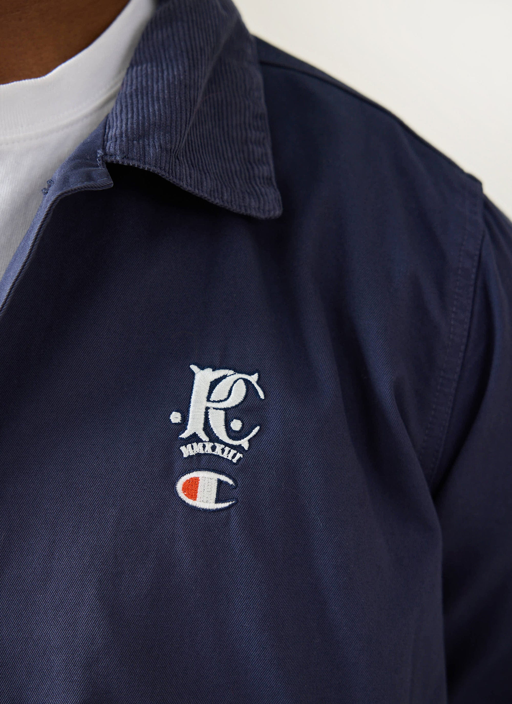 A close-up of the collaborative embroidery on the chest of the navy Champion x Percival Cord Collar Jacket.