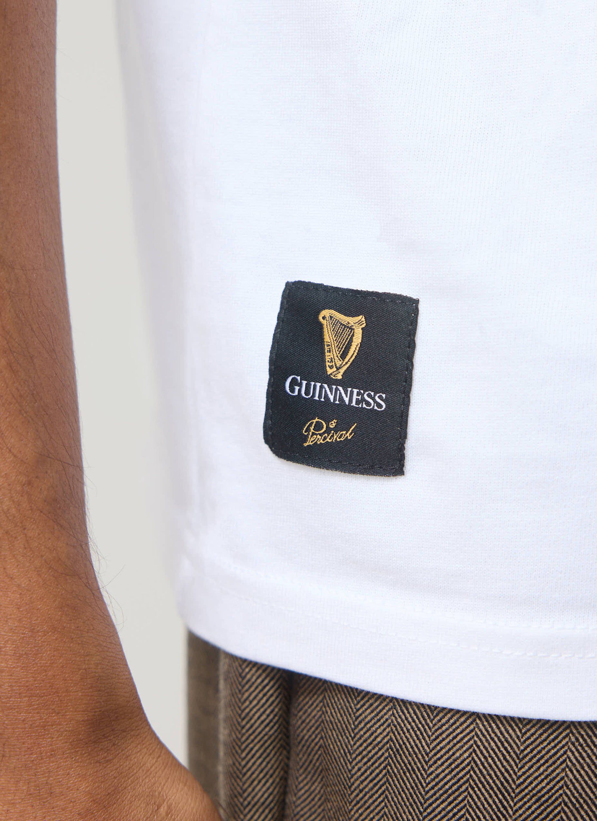 Close-up of the Guinness x Percival collaboration label, with gold embroidery, on the hem of the white Stout T Shirt.