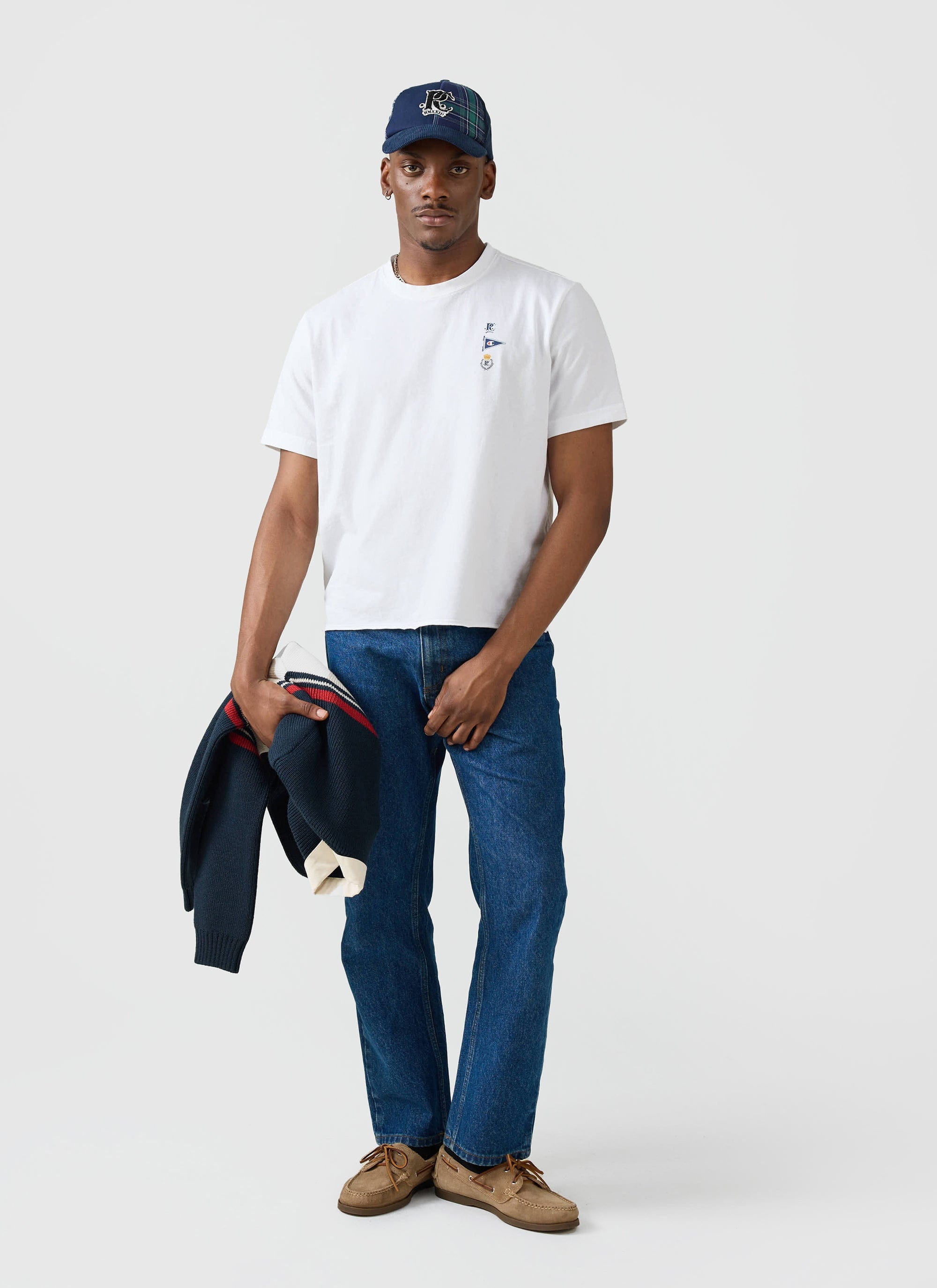 A model wears the white Champion x Percival Flag T-shirt with blue jeans and a cap, holding a navy jumper.