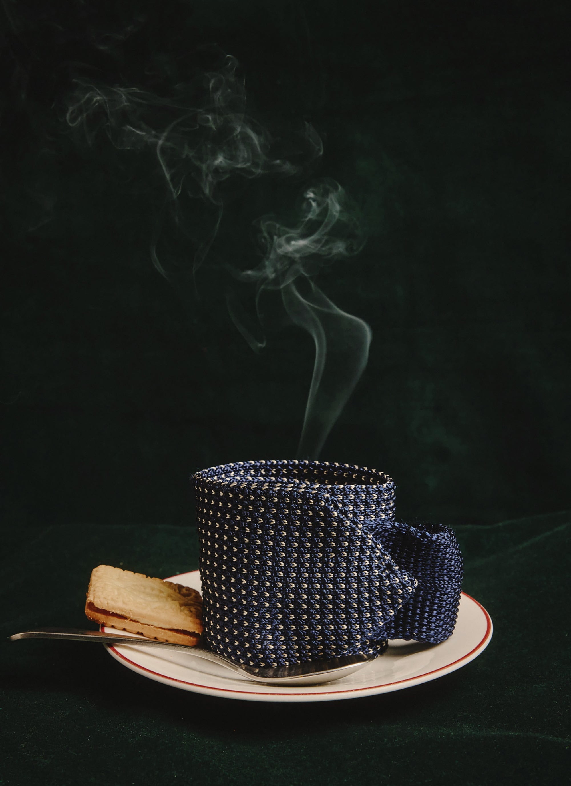 A creative still life of the Percival blue Birdseye Silk Tie, shaped into a steaming mug on a saucer with a biscuit.