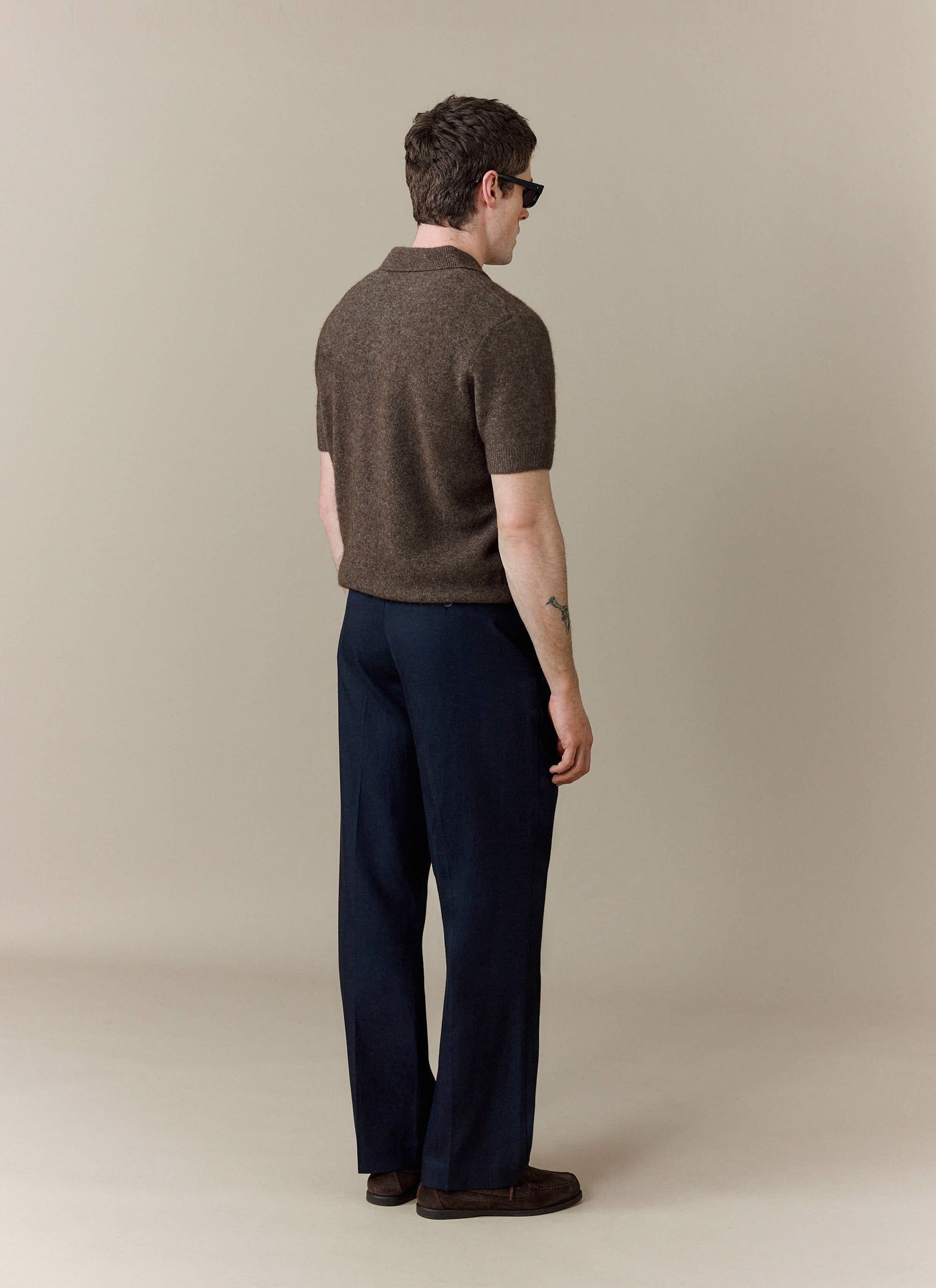 A model seen from the back wears the Percival Collins Boxy Knitted Polo in Espresso, styled with smart navy trousers.