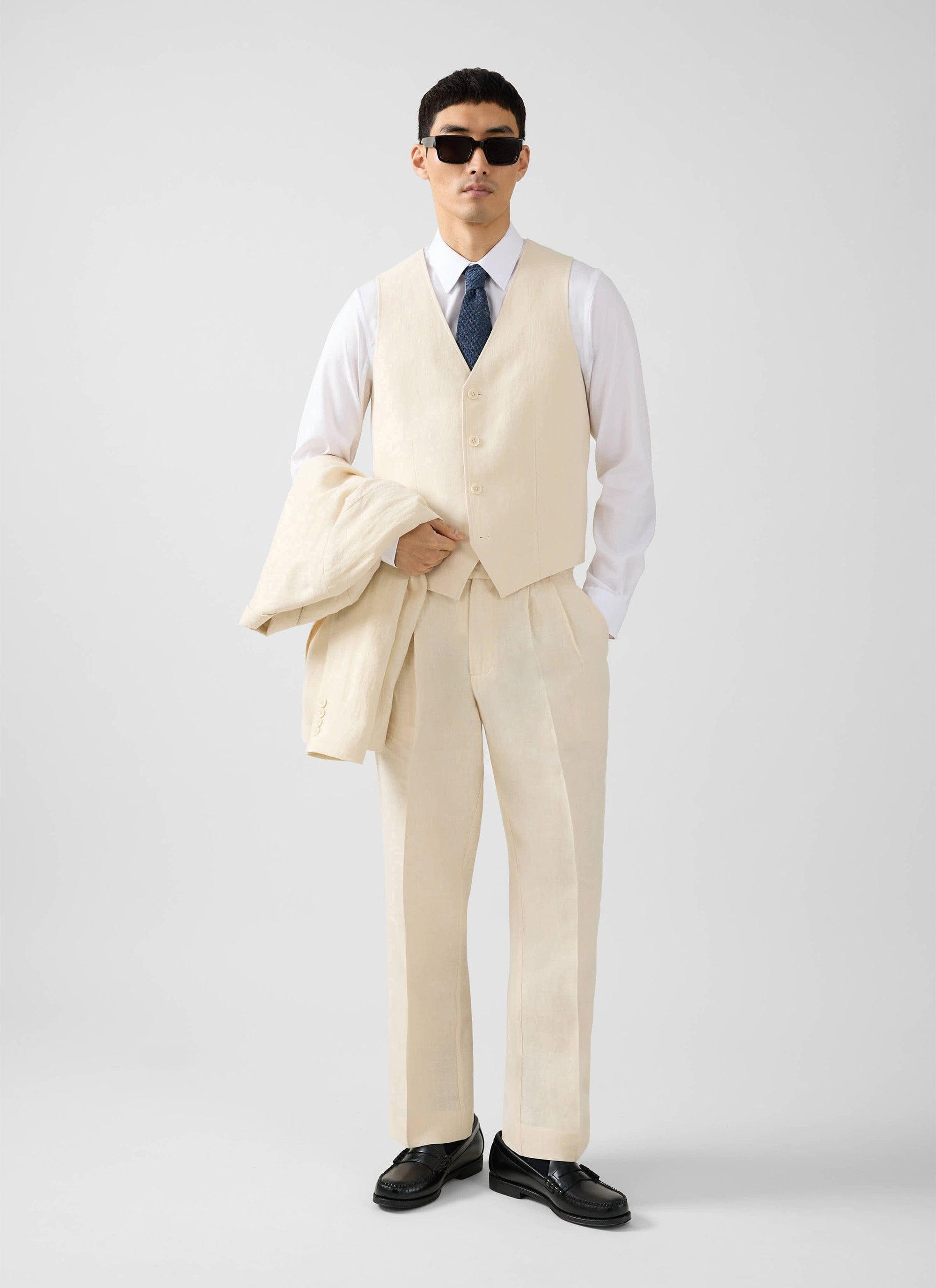 A model wears the Percival cream linen waistcoat with matching trousers, a white shirt, and blue tie, holding the jacket.