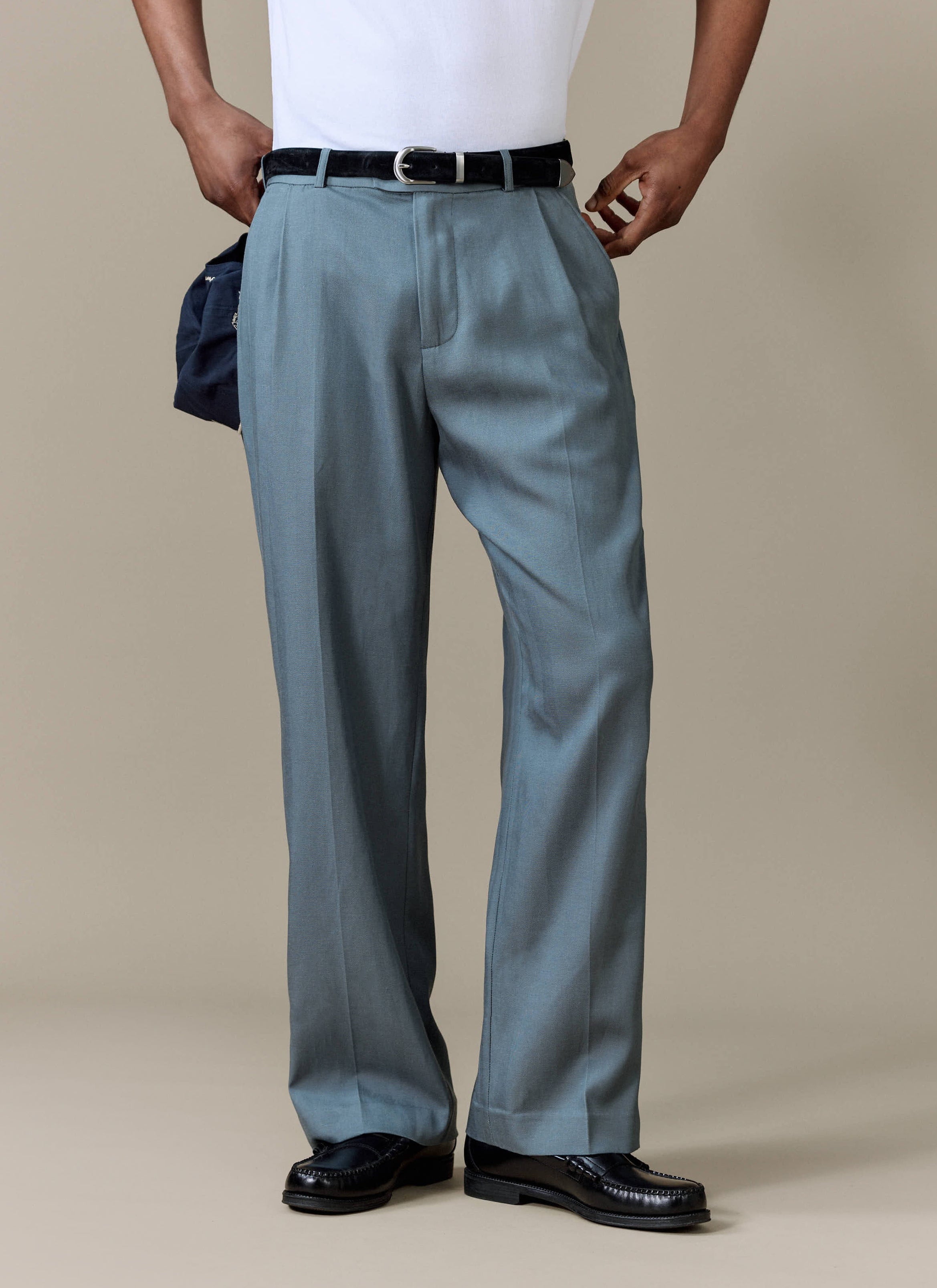 A model wears the slate blue Pleated Leg Trousers from Percival, styled with a white tee, belt, and black loafers.