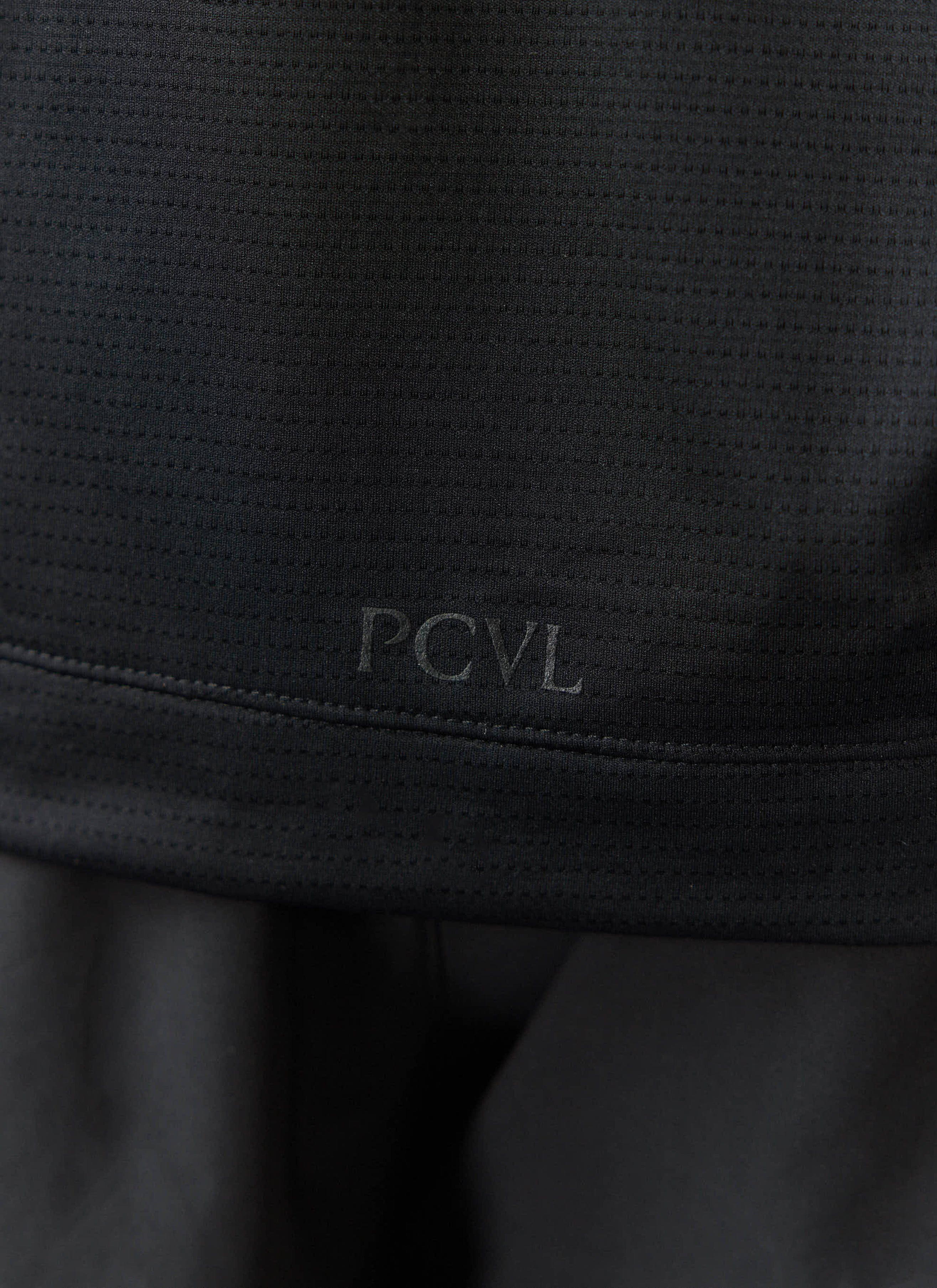 A close-up of the tonal PCVL logo on the hem of the black Performance Top by Percival, showing the breathable fabric texture.