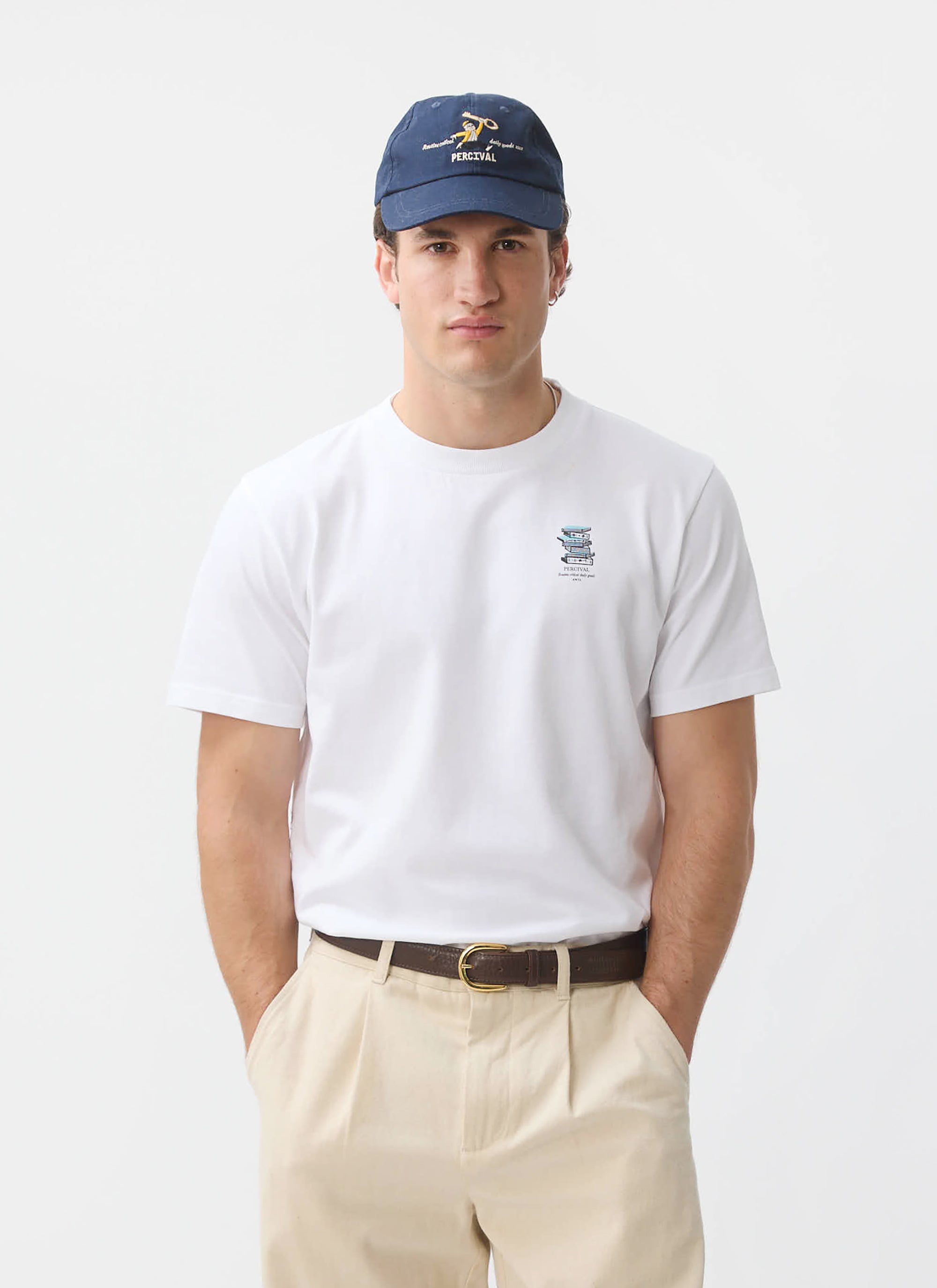 A model wears the white Duke T Shirt from Percival, featuring a book graphic, styled with beige trousers and a blue cap.