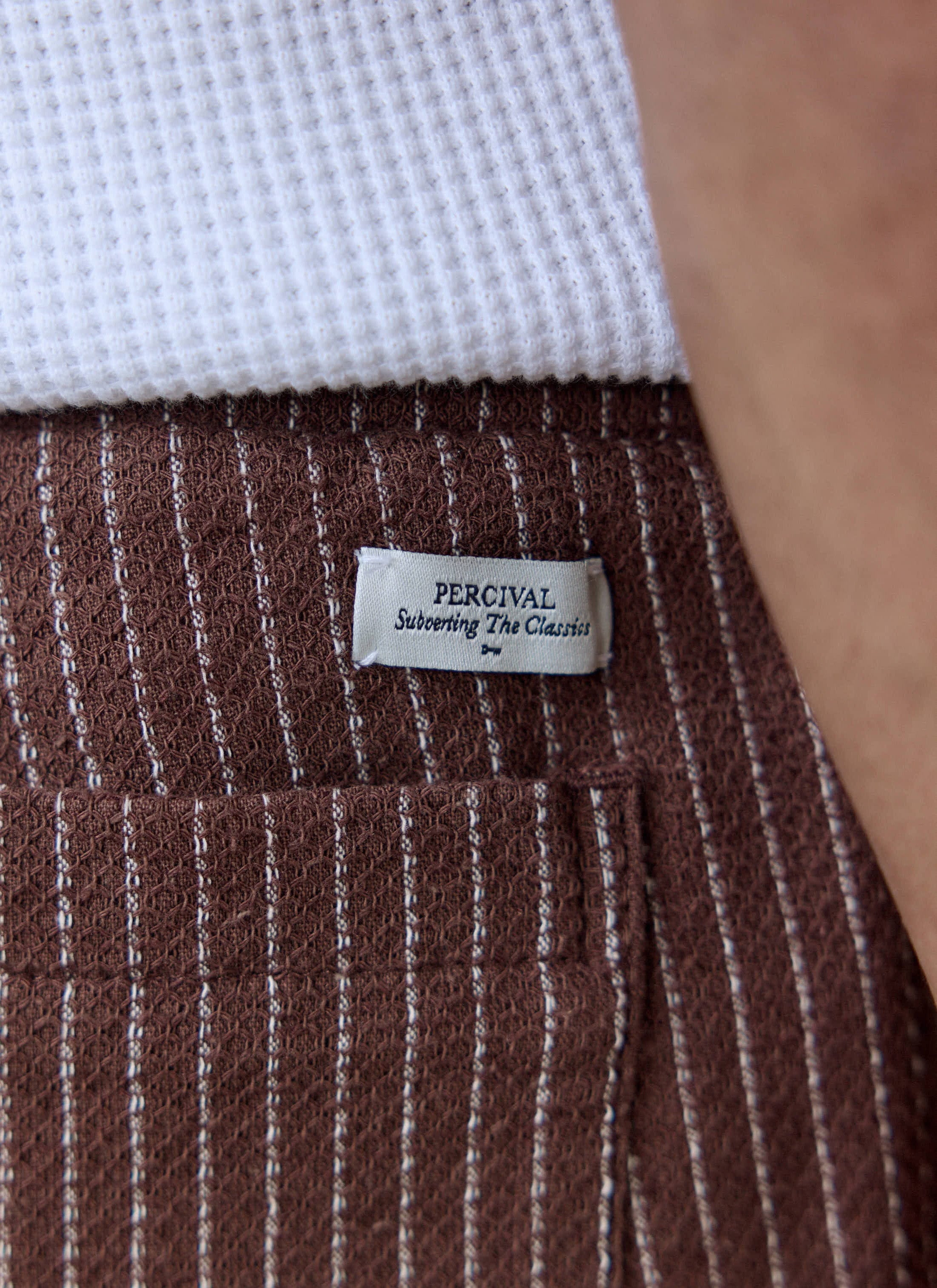 Close-up of the brown linen Sorrento Shorts from Percival, showing the subtle stripe and "Subverting The Classics" label.