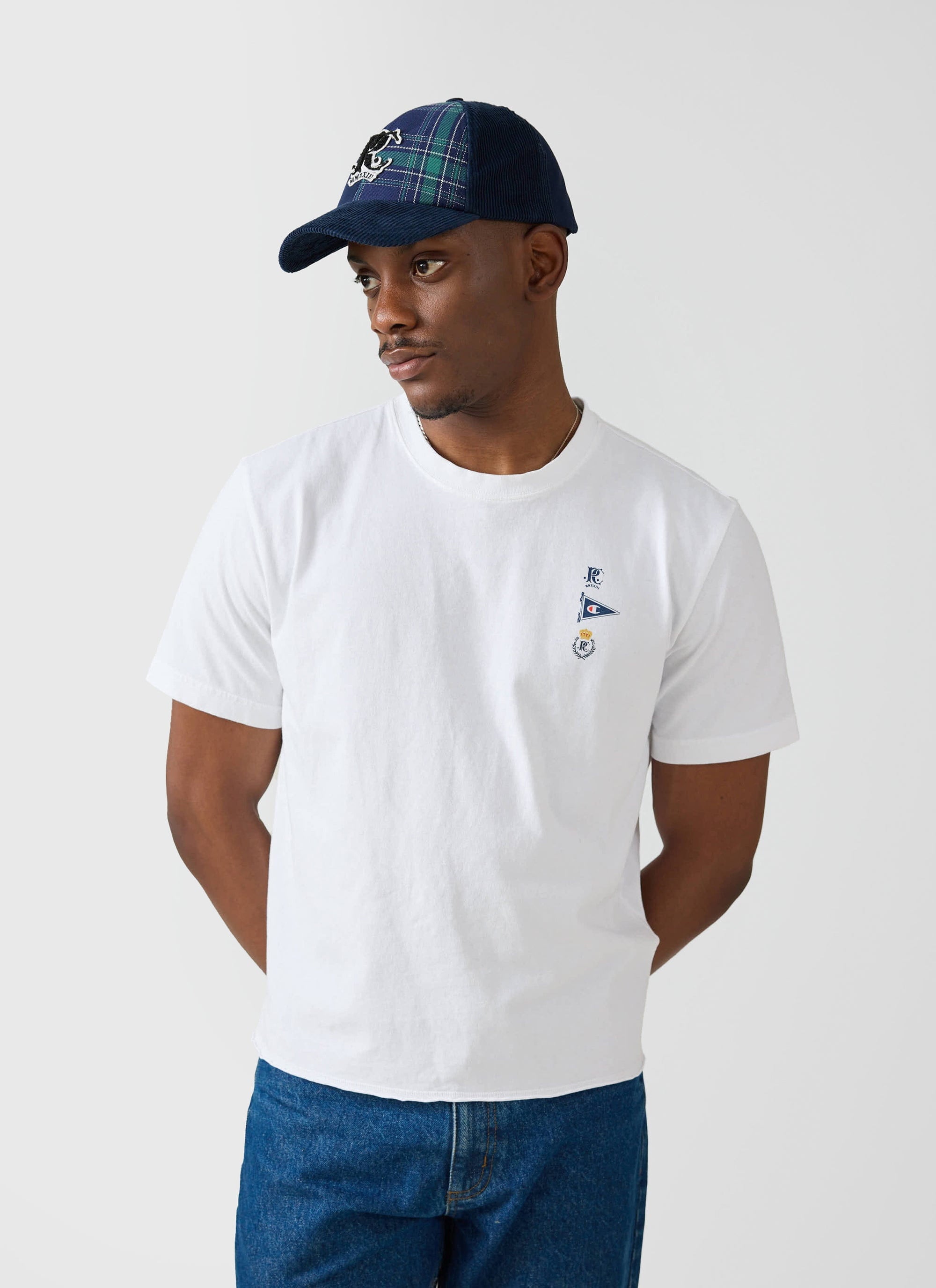 A model wears the white Flag Raw Hem T Shirt from the Champion x Percival collaboration, styled with a blue tartan cap.