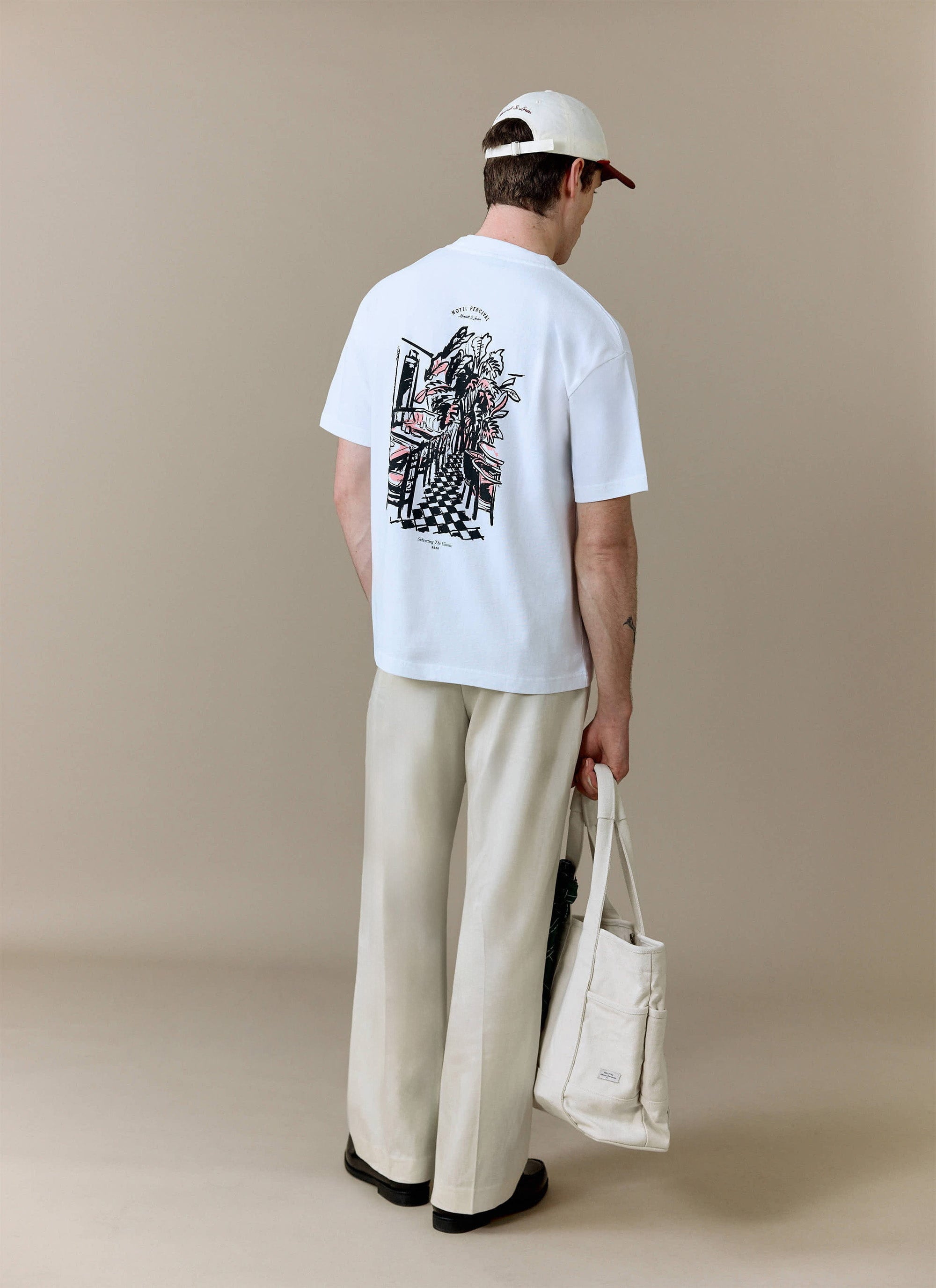 A model, seen from behind, wears the white Hotel Bistro Oversized T-Shirt from Percival with cream trousers and a tote bag.