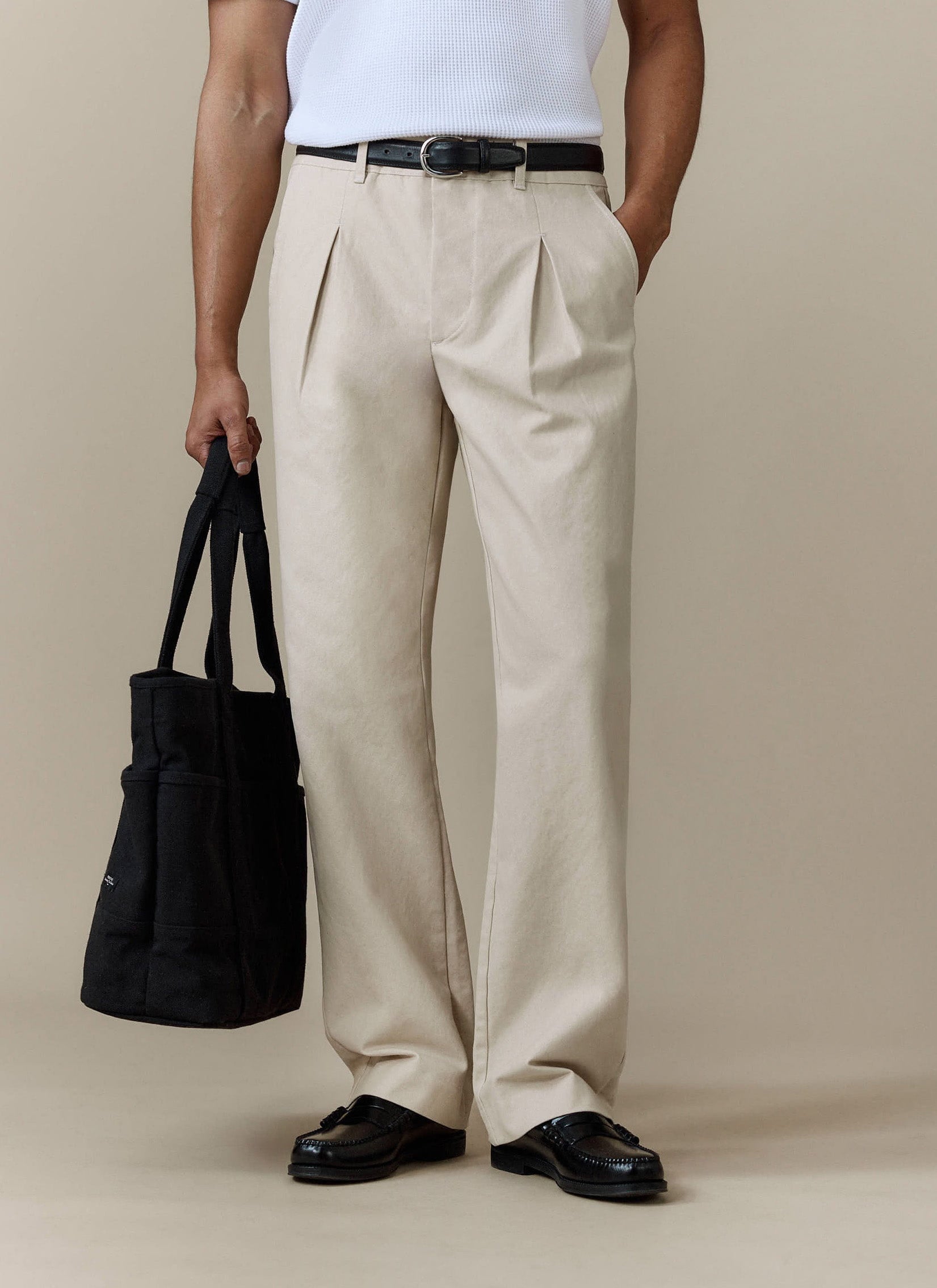 A model wears the Percival Seb Inverted Pleat Trousers in ecru, styled with a white top, black loafers, and a tote bag.