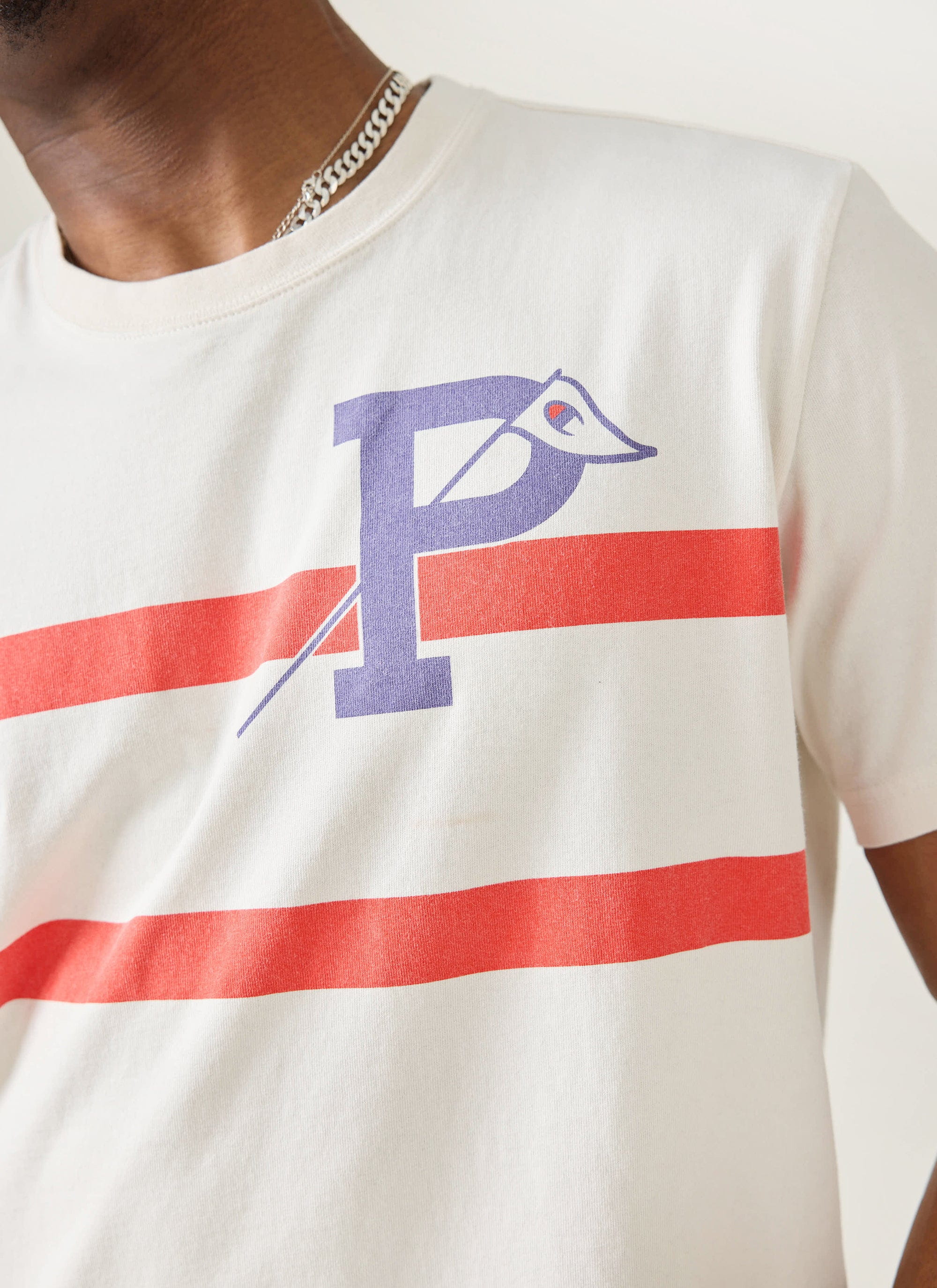 Close up of the Champion x Percival Flag Stripe T-shirt in ecru, showing the purple P-flag logo and red stripe chest detail.