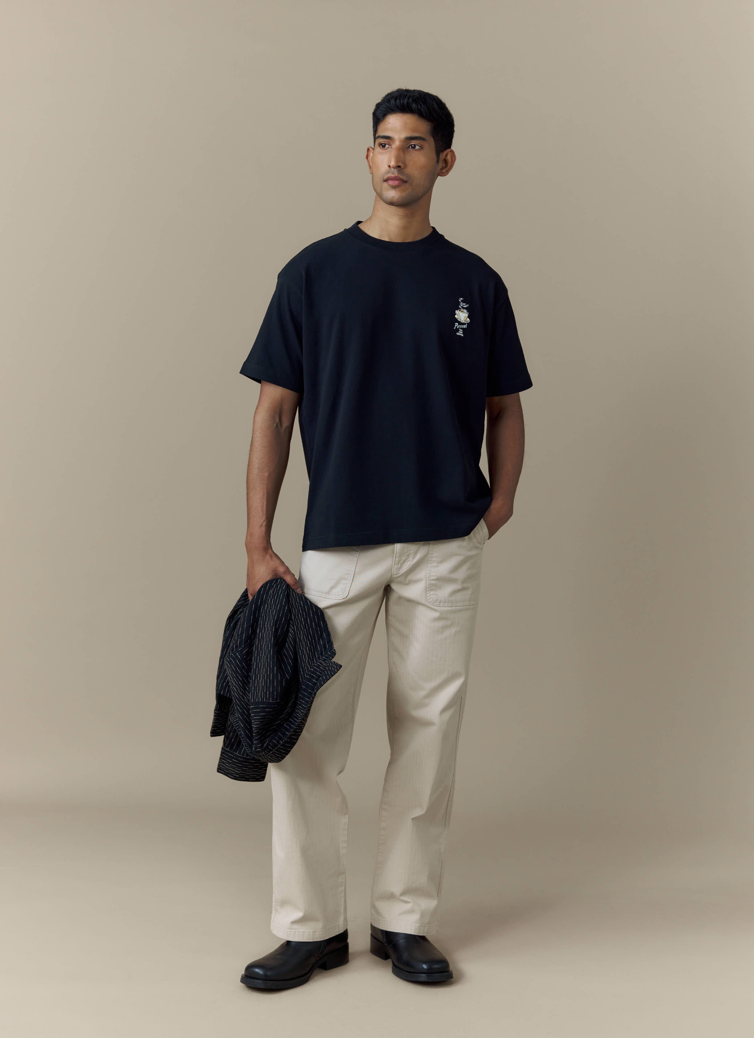 A man models the Percival Grill Oversized T Shirt in black, paired with off-white trousers and holding a jacket.