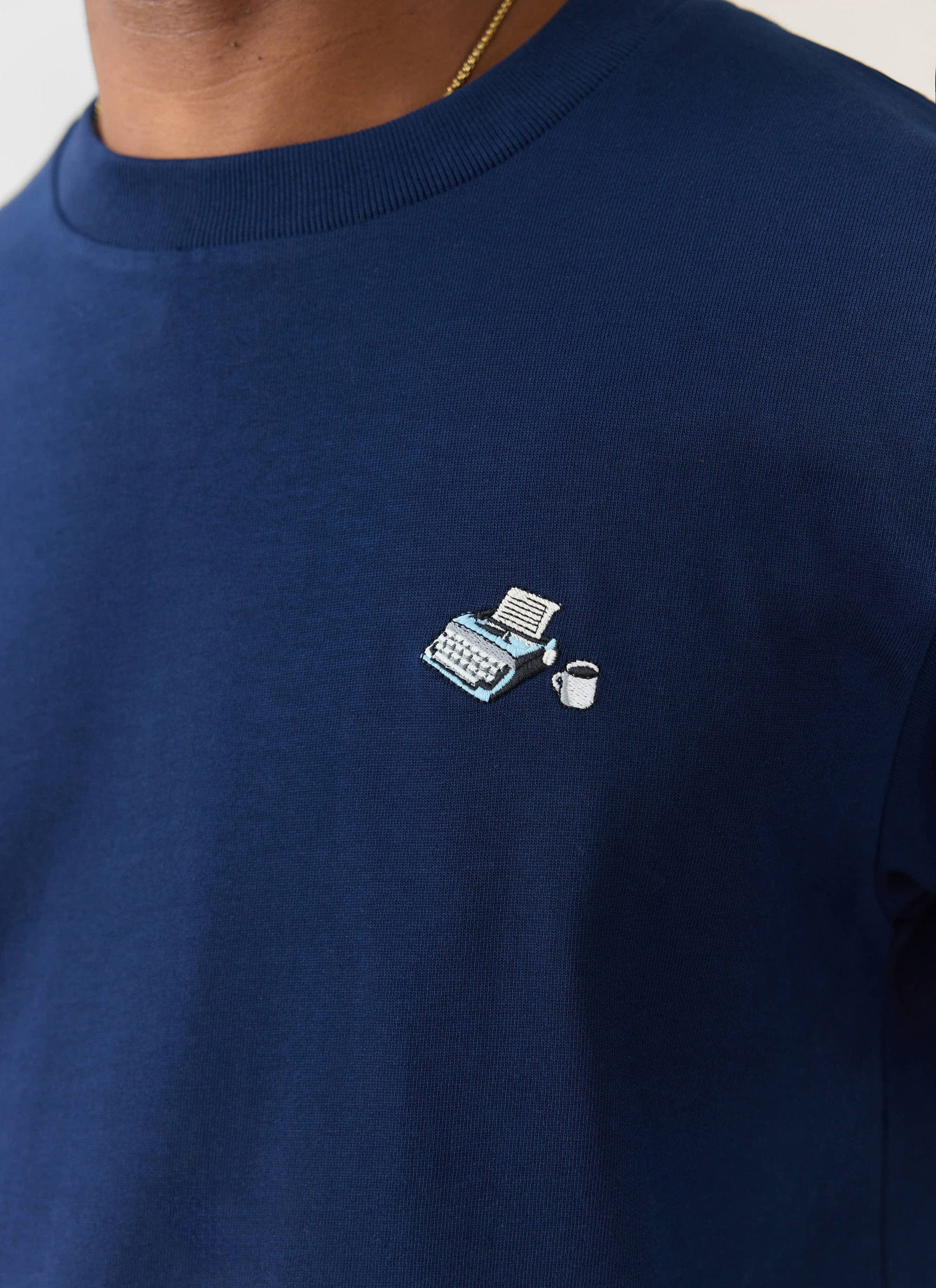 A close-up of the Percival Typewriter Embroidered T-Shirt in navy, showing the embroidery of a typewriter and coffee cup.
