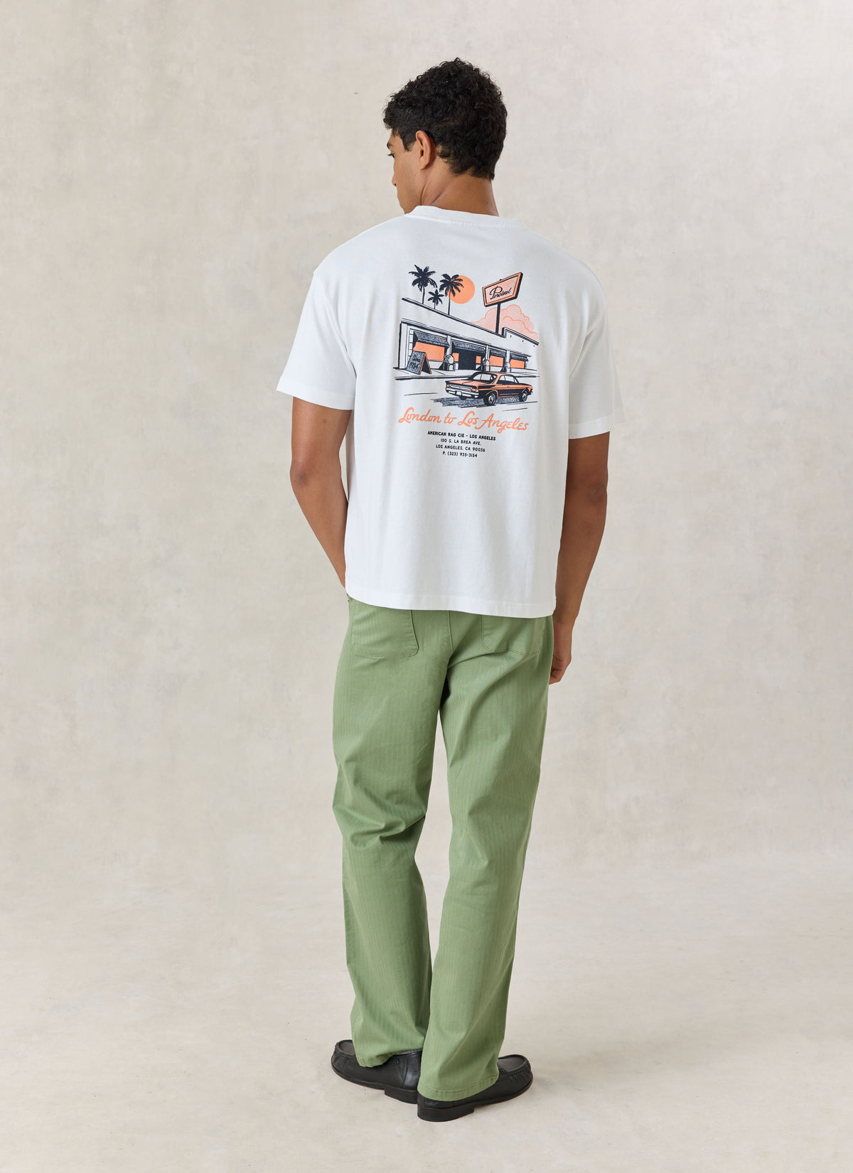 A model wearing the white American Rag x Percival oversized T-shirt, showing the back graphic print and green trousers.