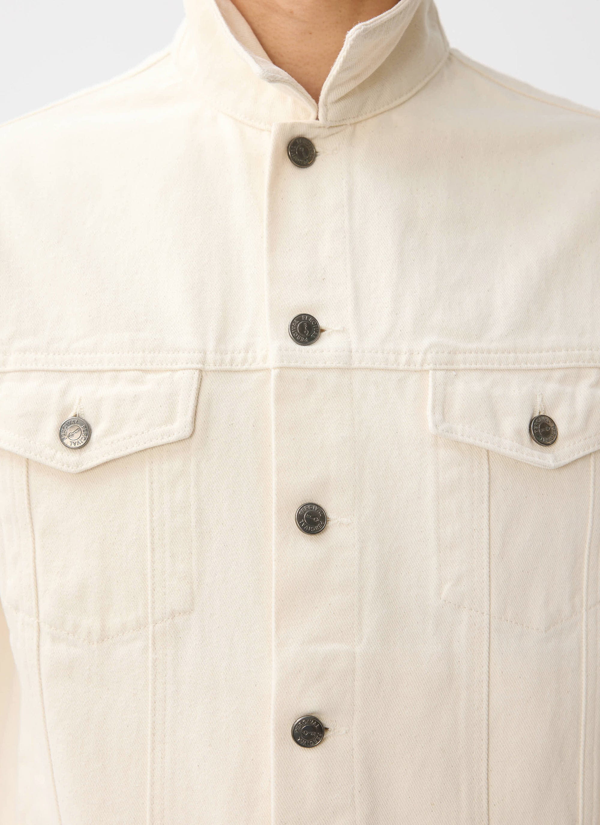 A close-up of the Percival Western Trucker Jacket in ecru denim, showing the branded buttons and chest pockets.