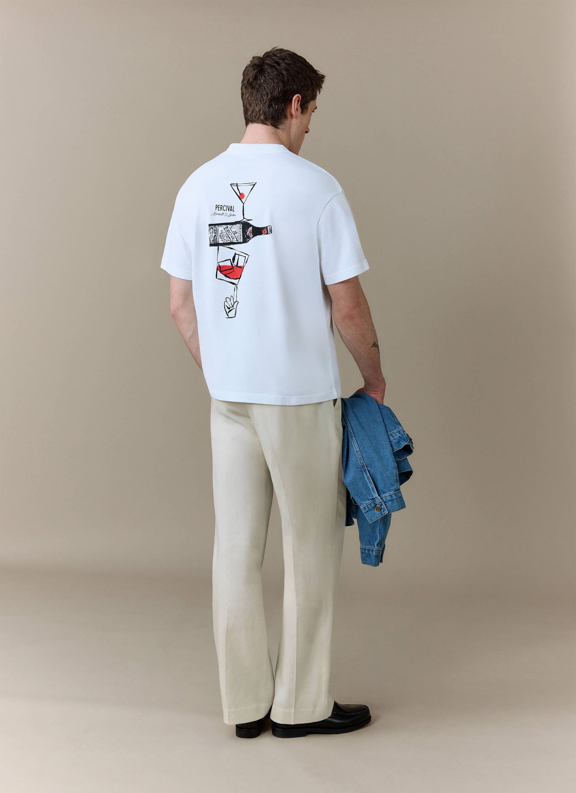 A model seen from the back wears the white Percival Perci & Rossi Martini tee with beige trousers, holding a denim jacket.