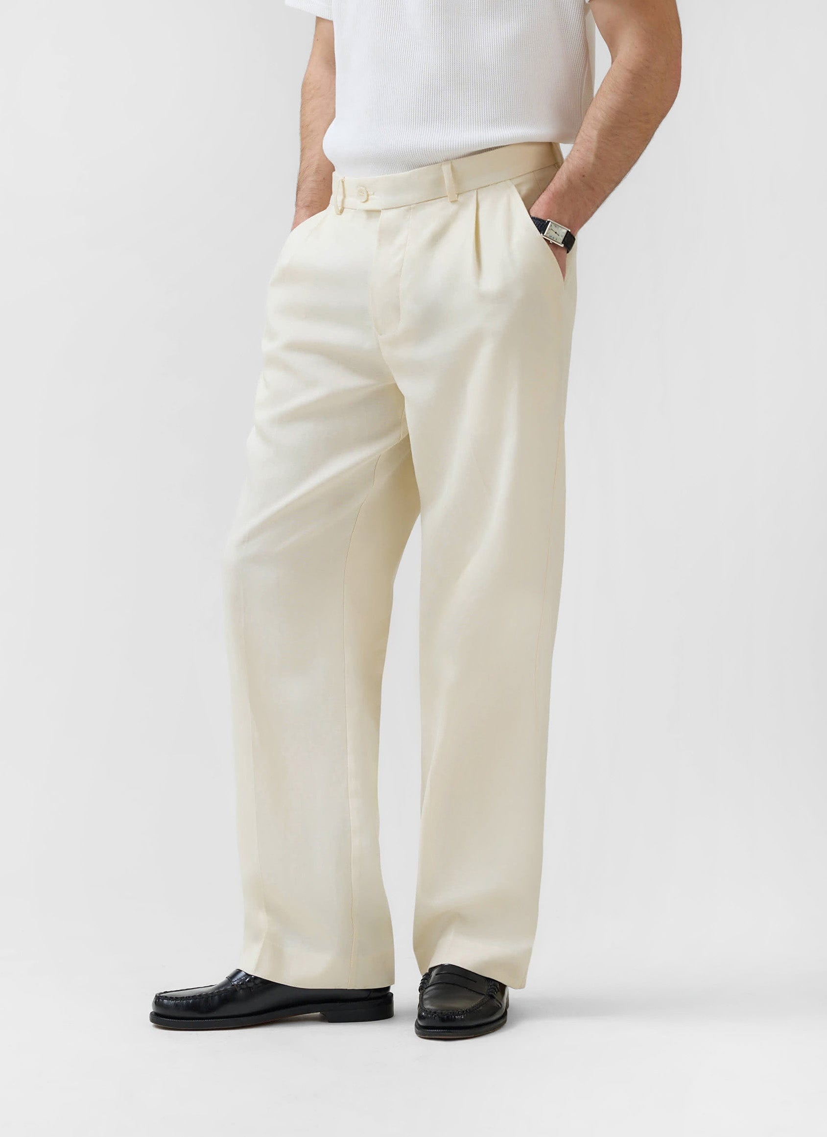 A man models the cream Pleated Tailored Trousers from Percival, styled with a white tee and black loafers.