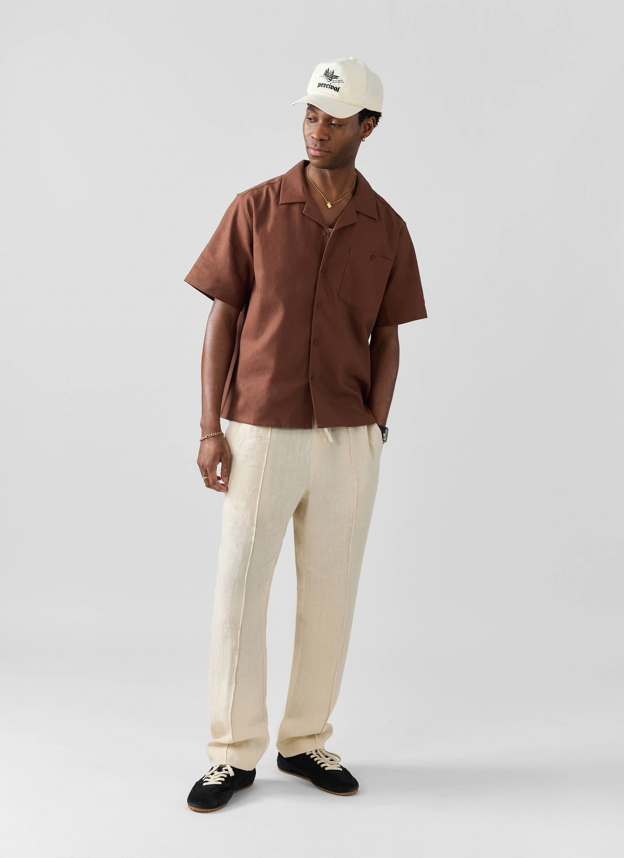A model styles the Percival Lorena Wide Leg Trousers in cream linen with a brown shirt and a cap in a full-length shot.