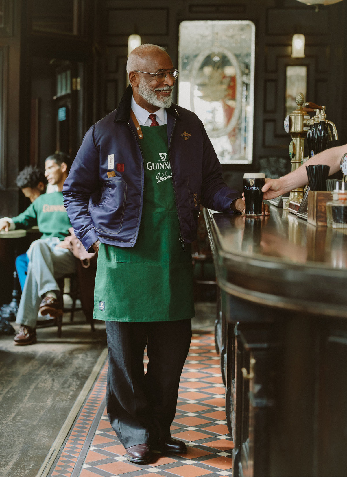 A model wearing the Guinness x Percival forest green apron at a pub bar, receiving a pint of Guinness.