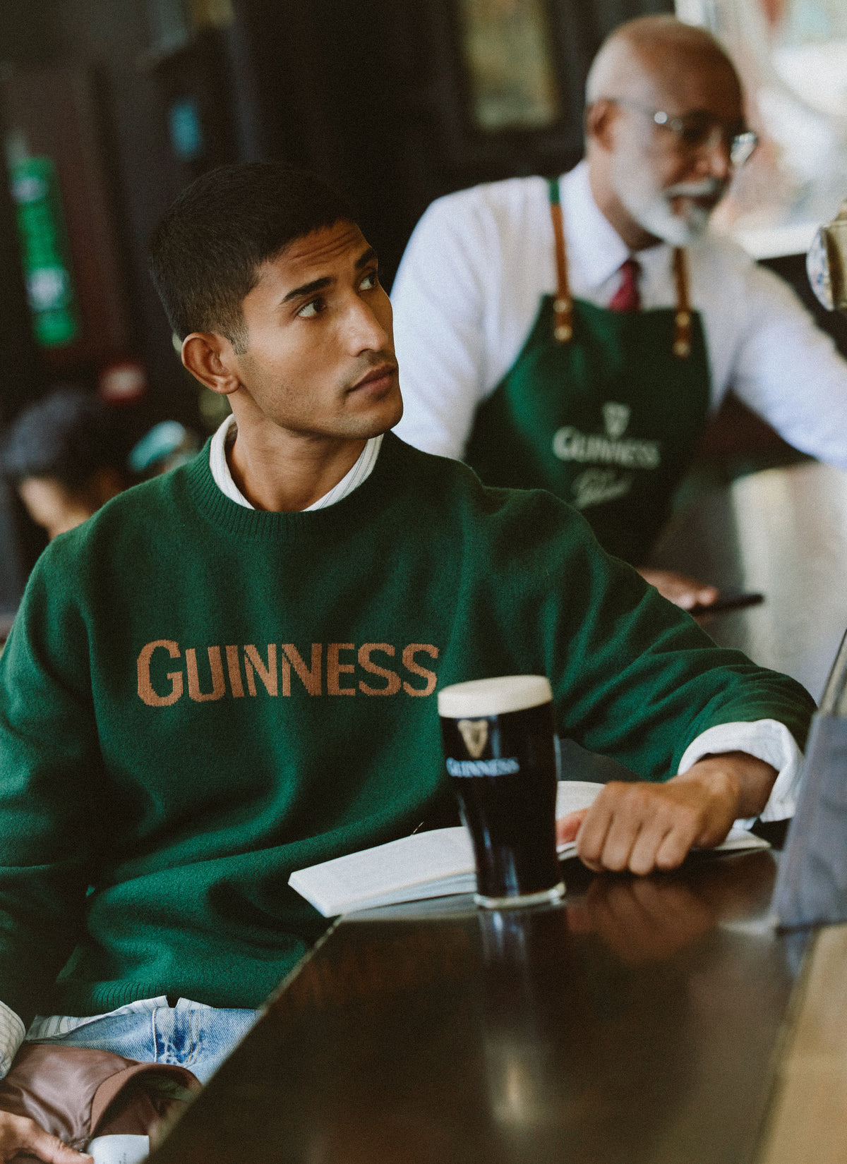 A model wearing the Guinness x Percival forest green wool jumper at a pub with a pint of Guinness.