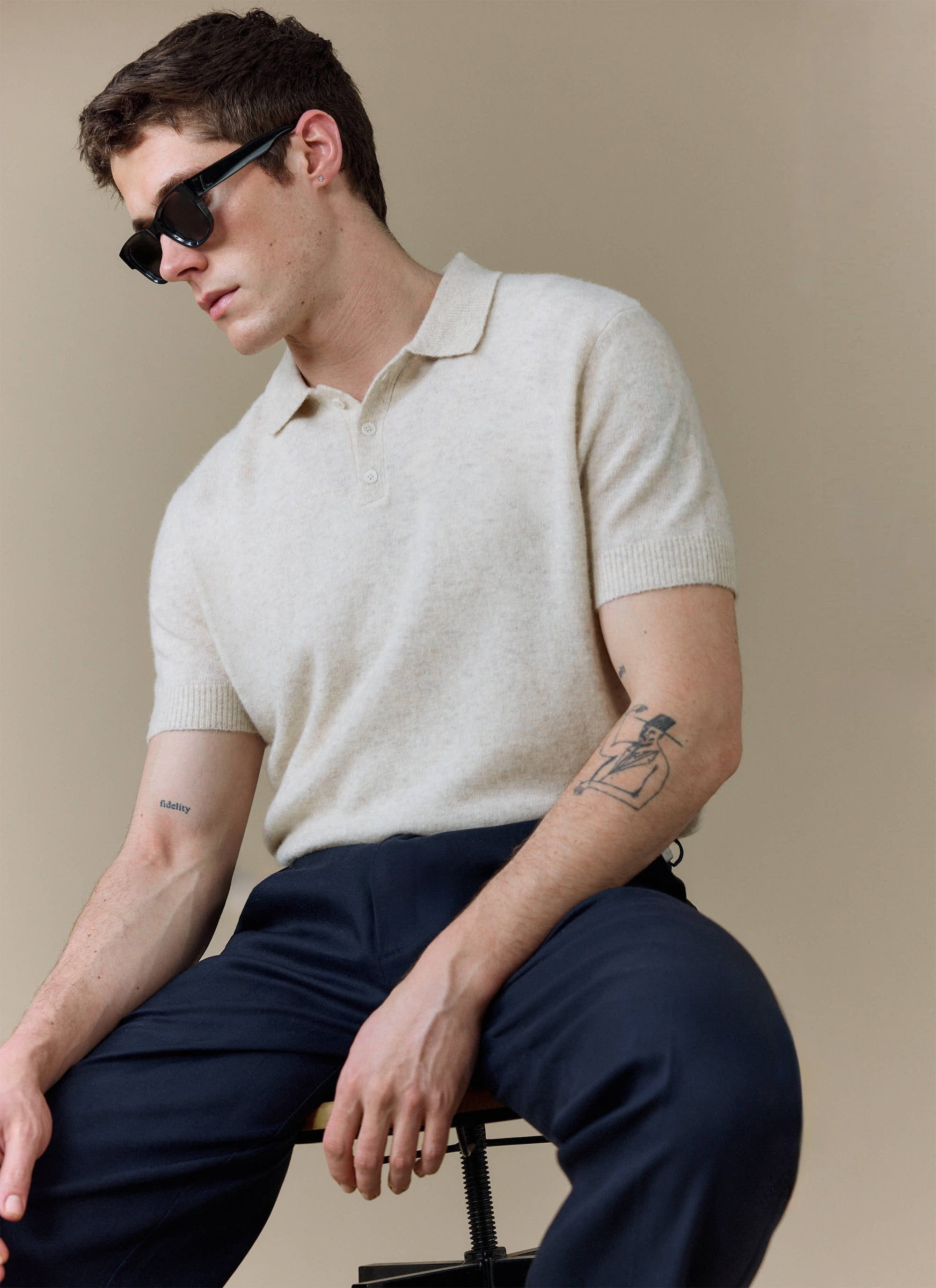A model sits wearing the Percival Collins Boxy Knitted Polo in ecru, styled with dark trousers and sunglasses.
