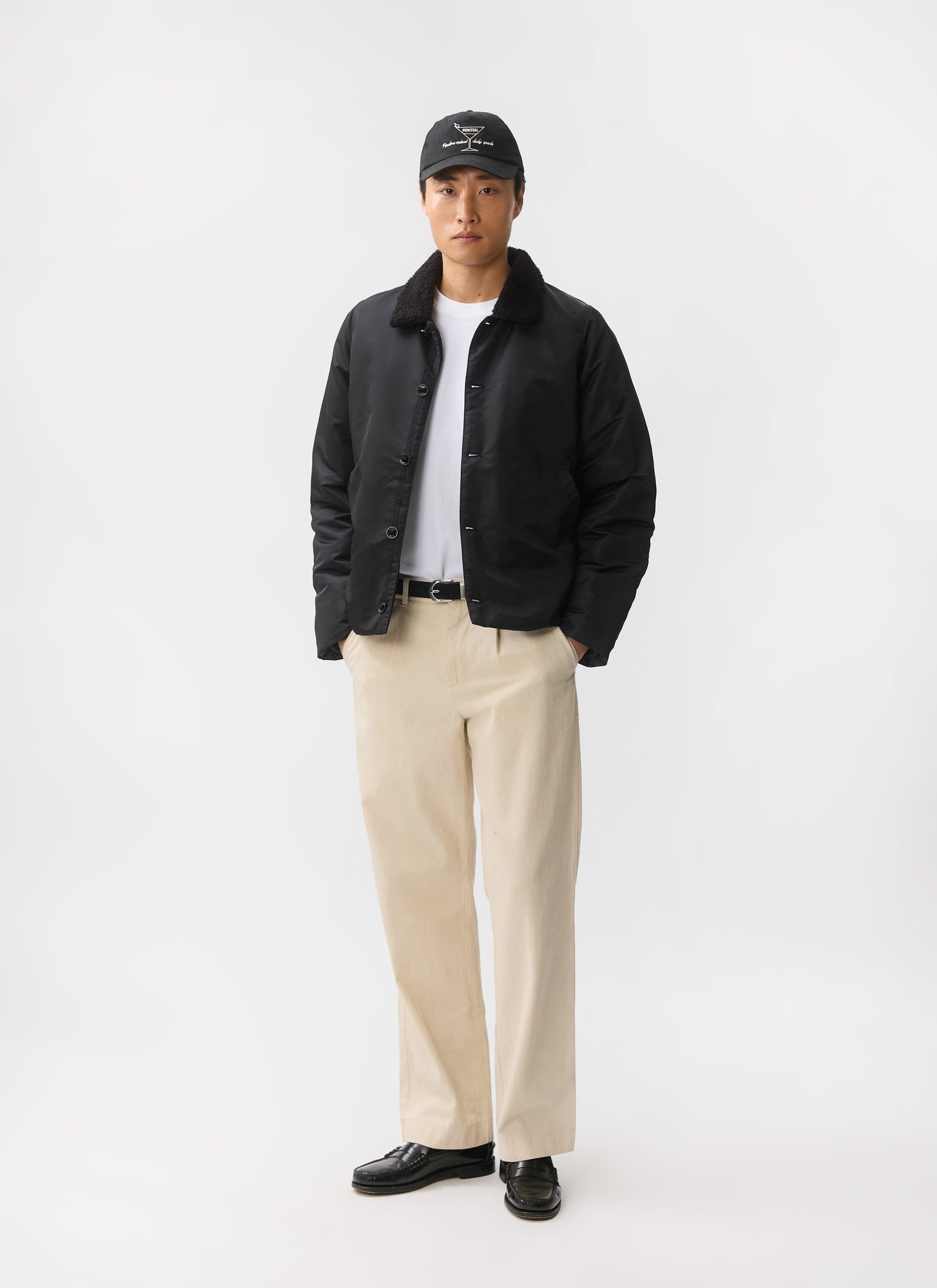A model wears the black Doppio Bomber Jacket from Percival, styled with a white t-shirt, beige trousers and a cap.