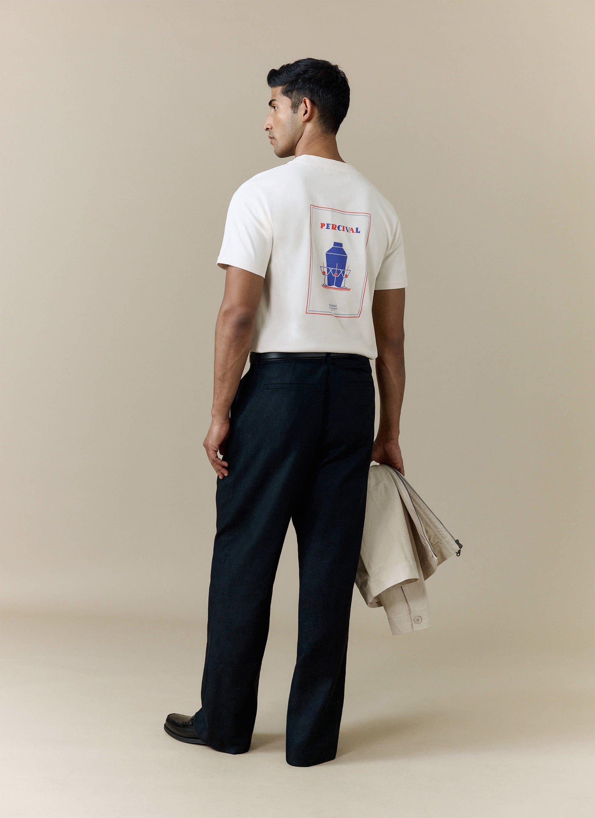 A model shows the back graphic of the ecru Mixologist Martini T Shirt from Percival, styled with dark trousers.