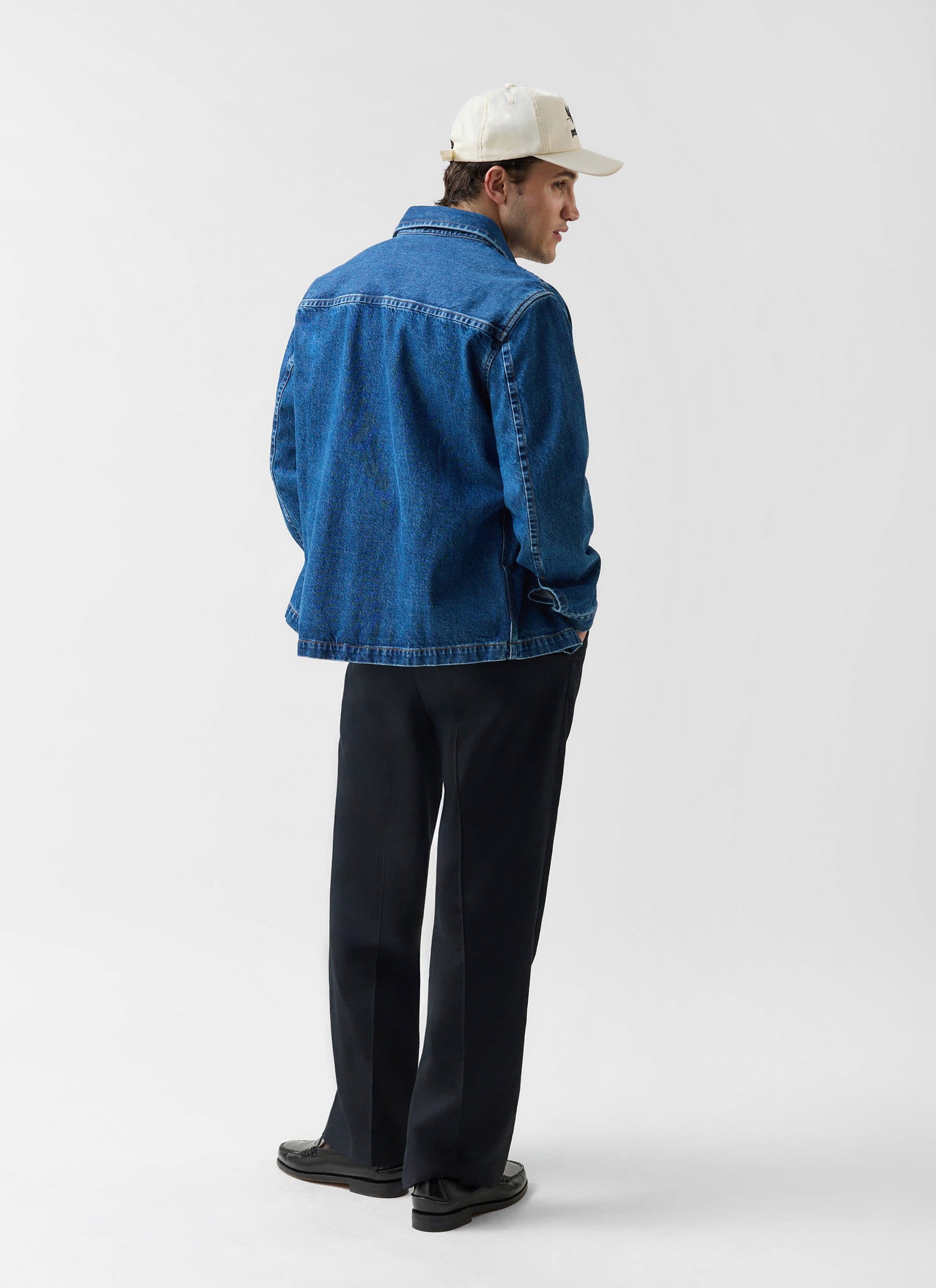 Rear view of a model wearing the Percival Denim Heavyweight Overshirt in mid-wash blue with black trousers and a white cap.