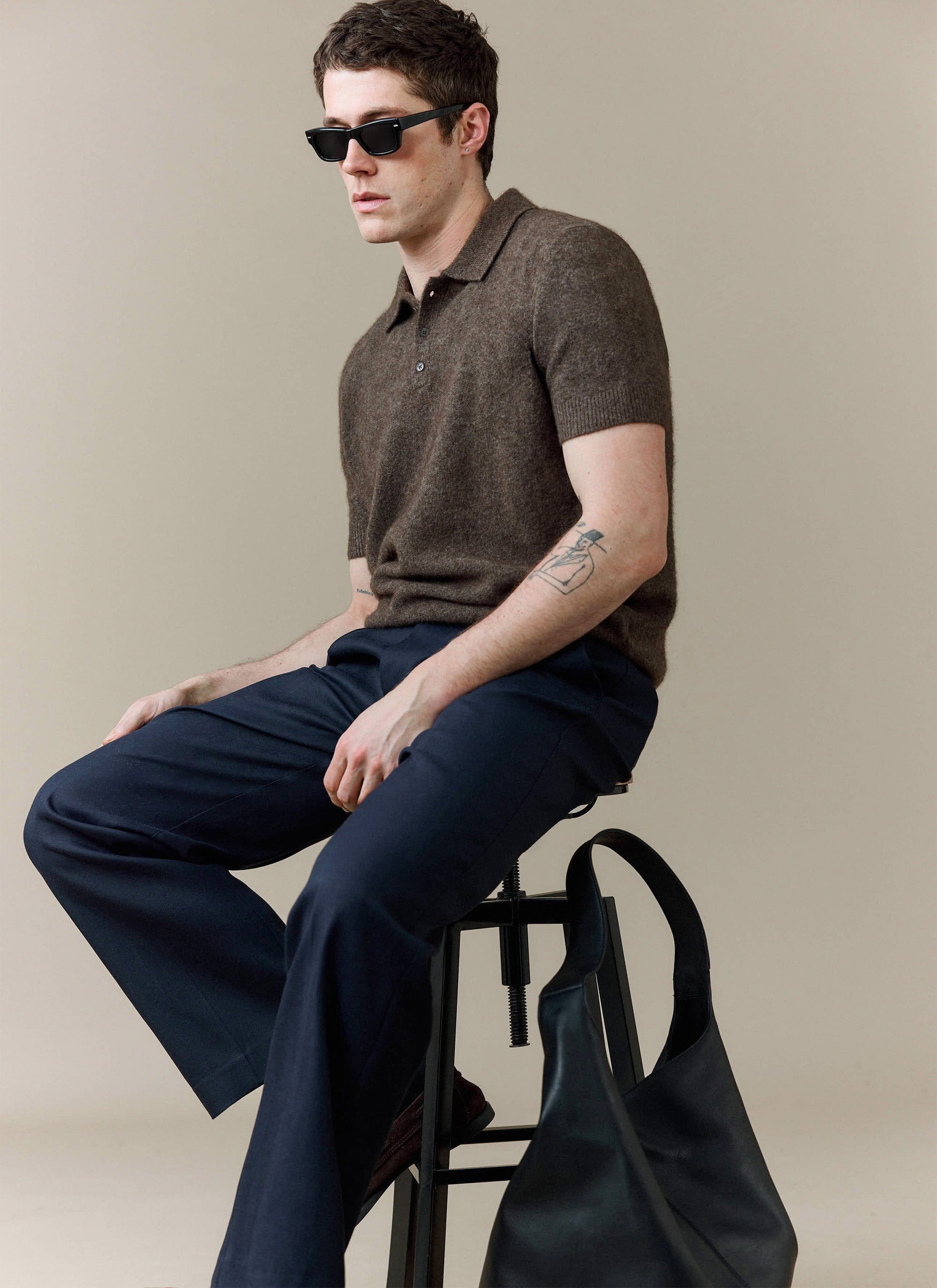 A man models the Percival Collins Boxy Knitted Polo in Espresso, styled with dark trousers and sunglasses.