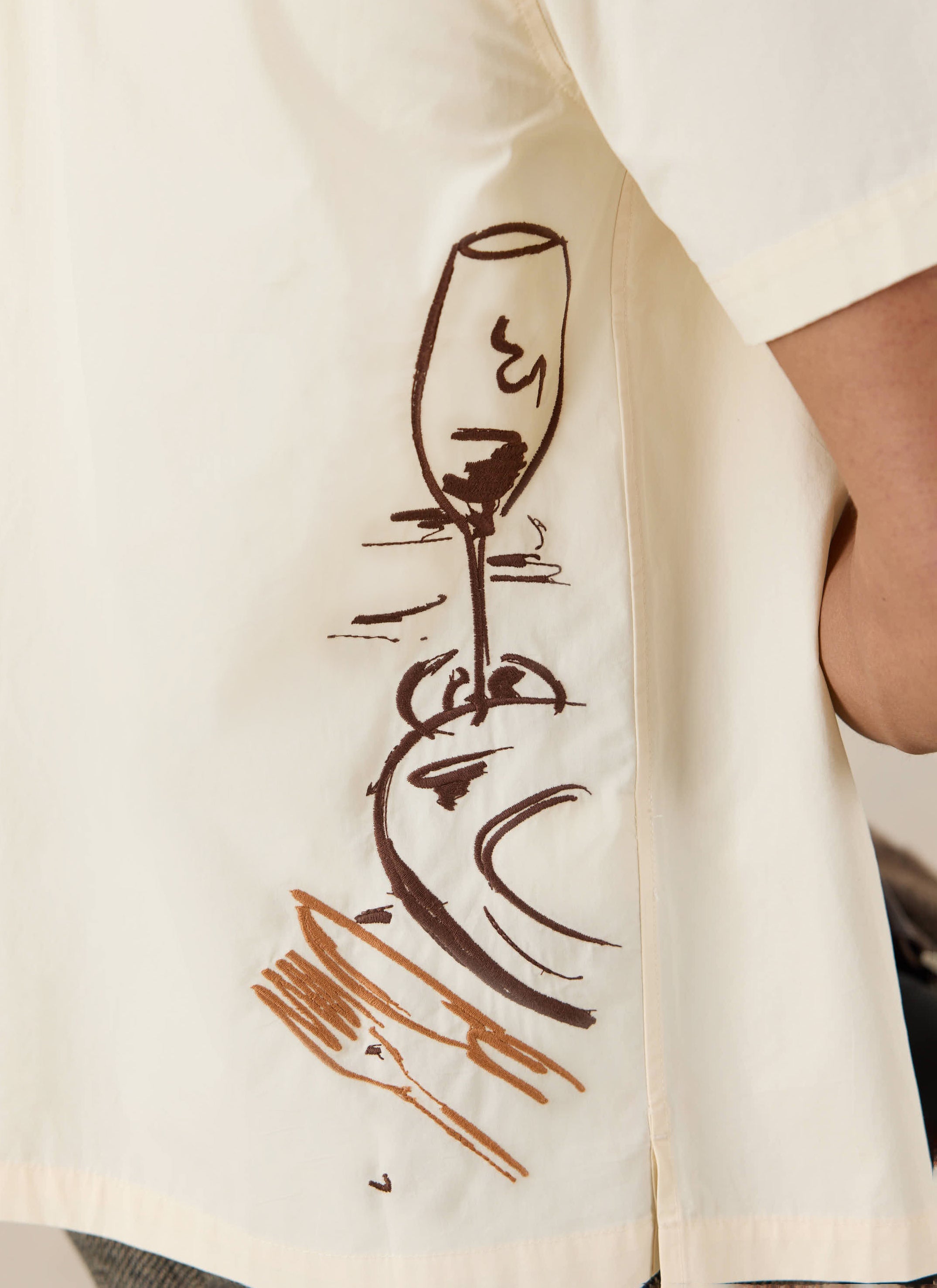Close-up of the cream cotton Percival Supper Club shirt, featuring a brown illustrative wine glass and plate graphic.