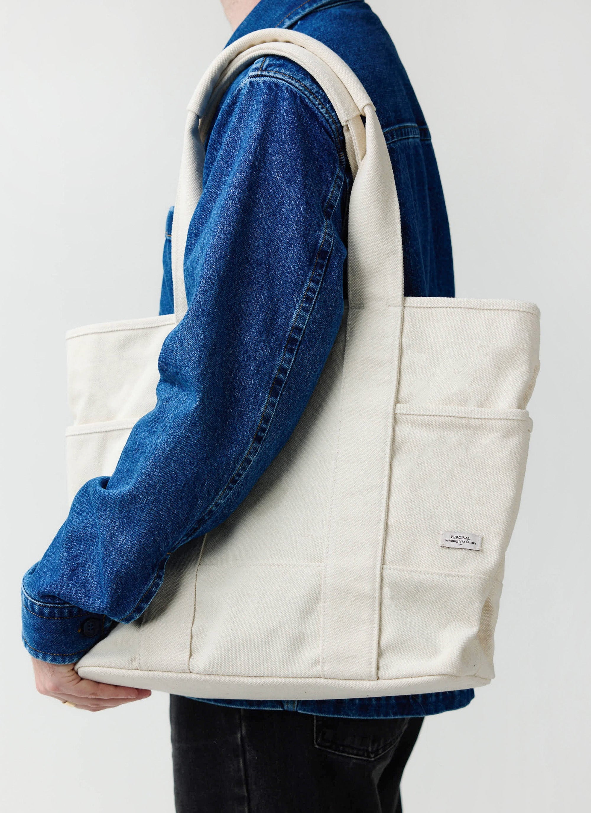A model wears the ecru cotton canvas tote bag from Percival over their shoulder, styled with a blue denim jacket.