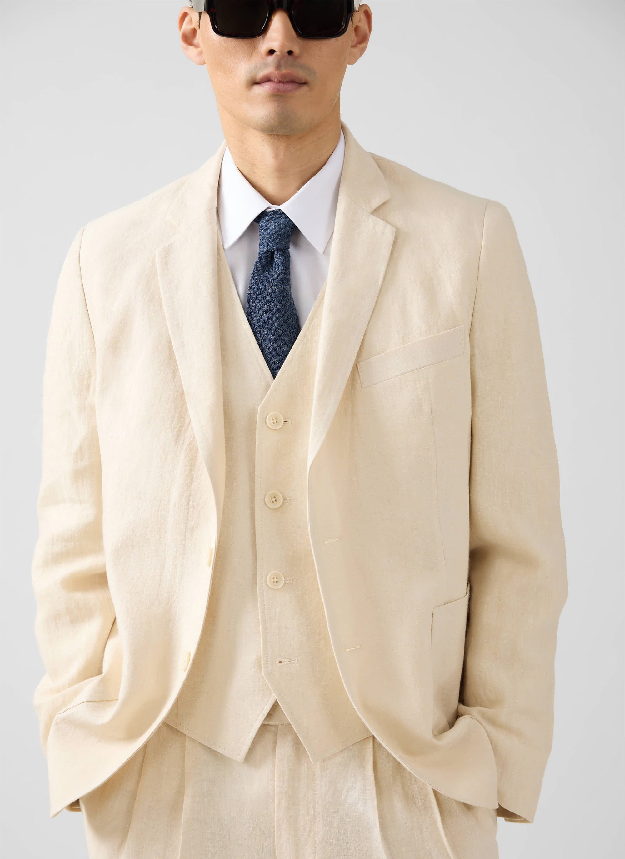 A model wears the Percival three-piece linen suit in cream, styled with a white shirt, blue tie and sunglasses.