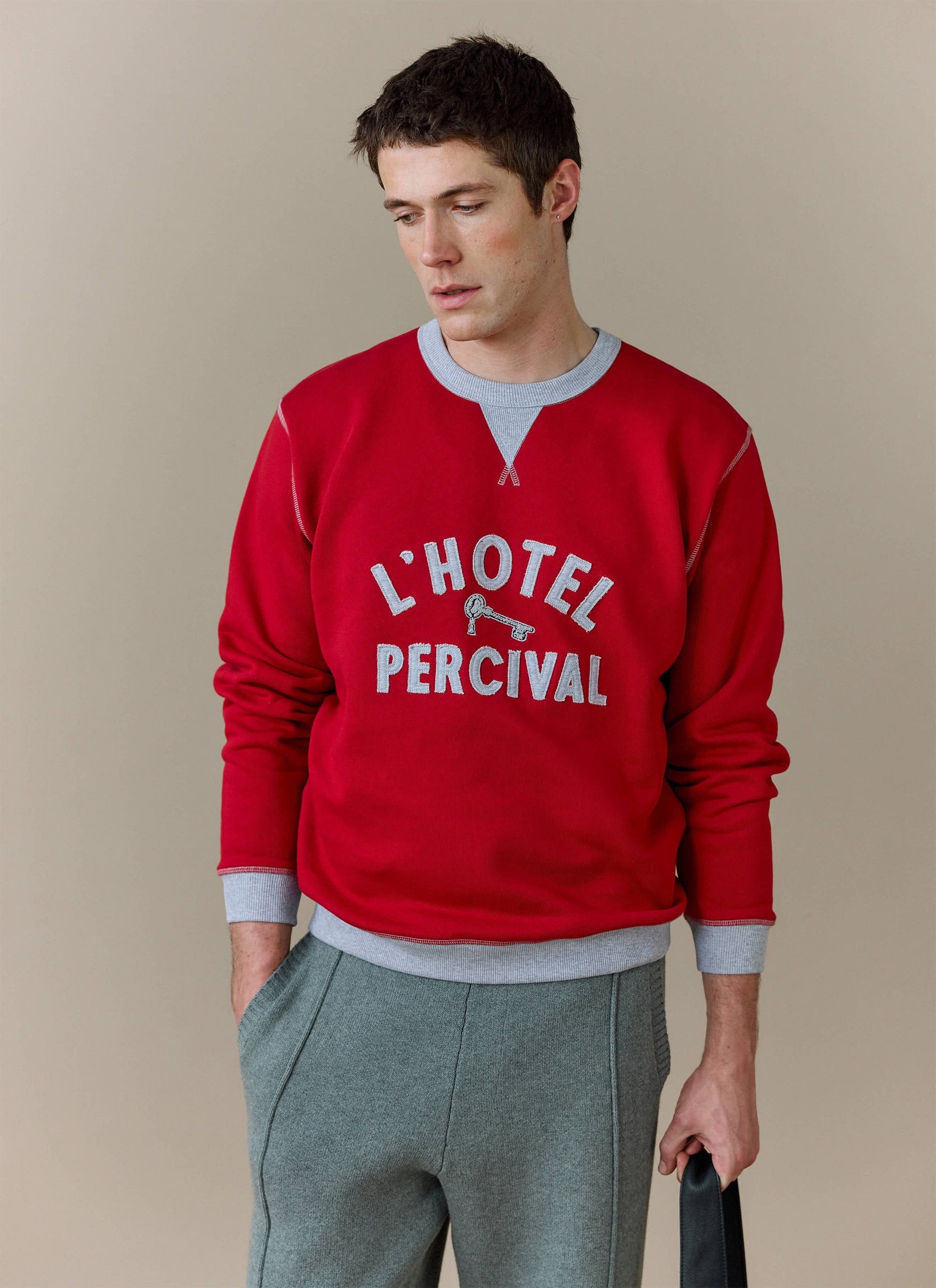 A man models the red Ronald Sweatshirt from Percival, featuring 'L'Hotel Percival' applique lettering, and grey trousers.