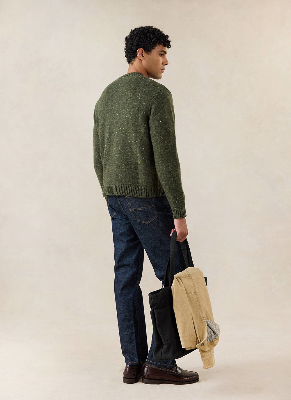 A model from the back wears the green Percival Rhodes jumper, styled with dark denim and a black tote bag.