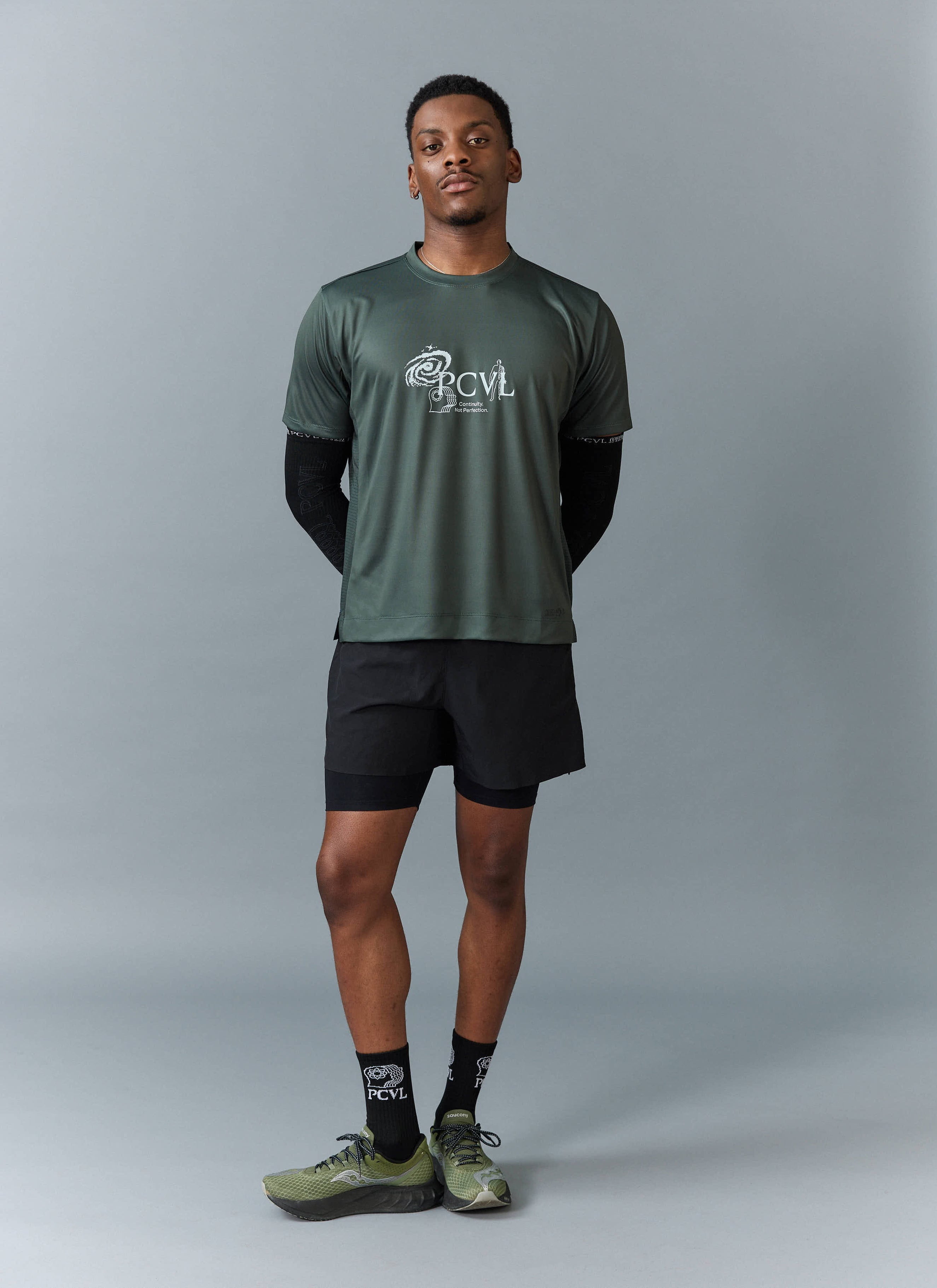 Full-length shot of a model wearing the Percival PCVL Printed Performance Top in forest with black shorts and trainers.