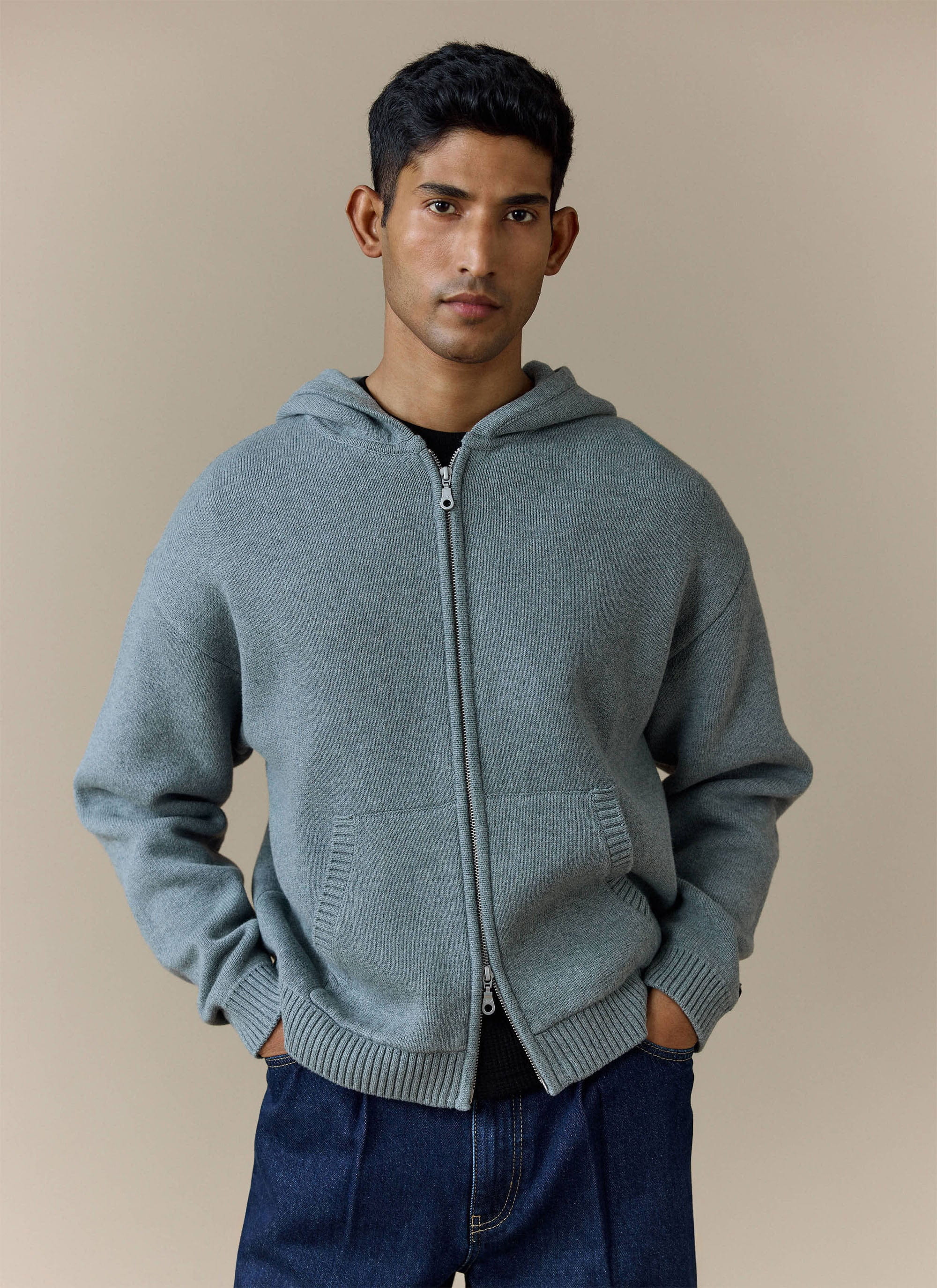 A man models the grey cotton Chronicle Knitted Hoodie from Percival, worn over a black t-shirt with dark jeans.