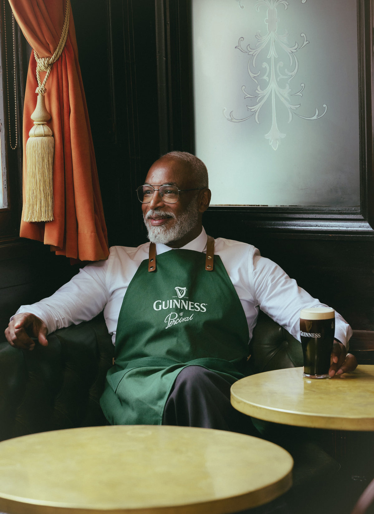 A model wearing the Guinness x Percival forest green apron while sitting in a pub with a pint of Guinness on the table.