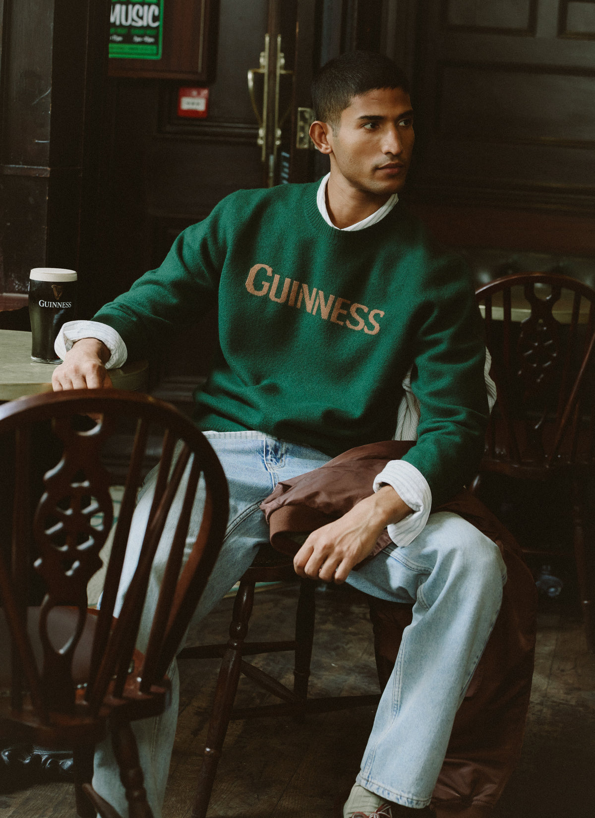 Model wearing the Guinness x Percival forest green merino wool jumper with jacquard chest lettering while seated in a pub.