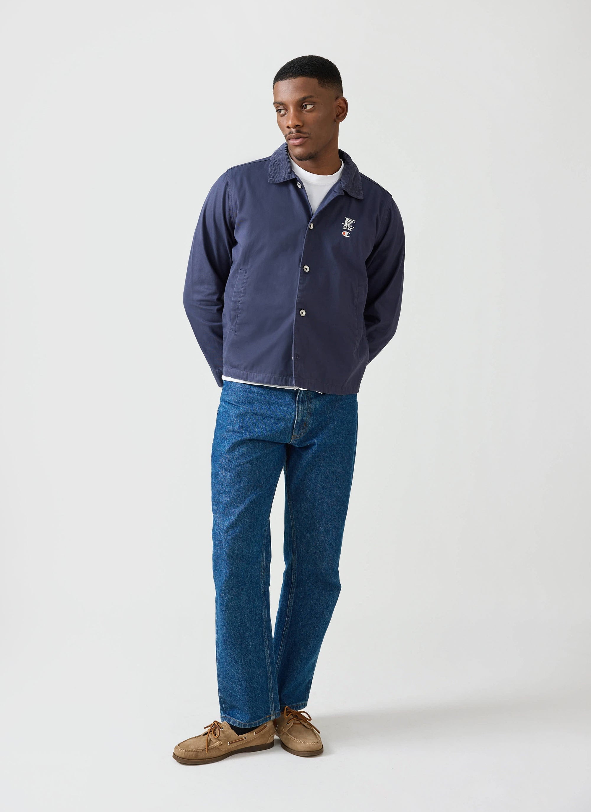 A man models the navy Champion x Percival Cord Collar Jacket with blue jeans and a white t-shirt in a studio.