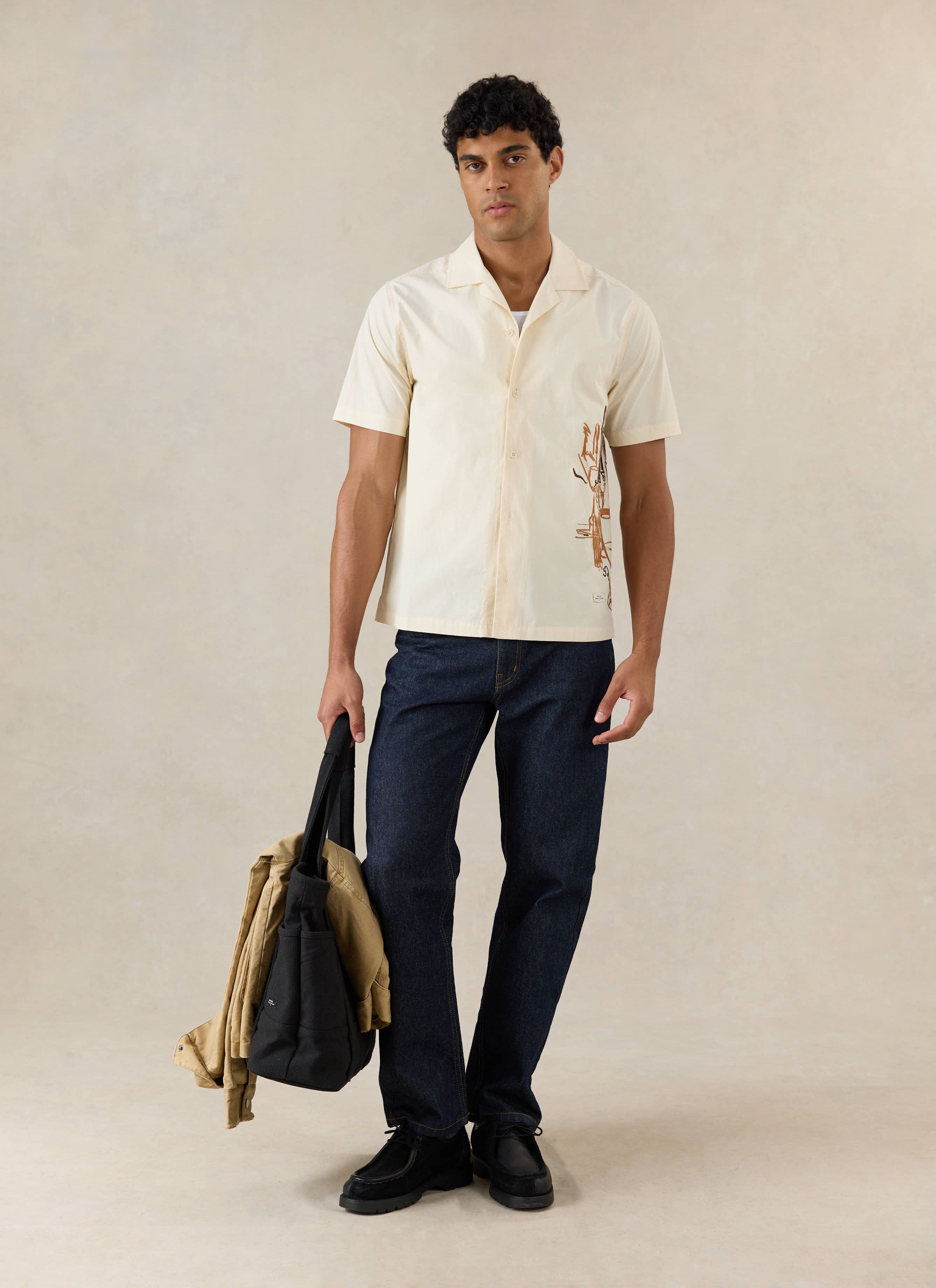 A model wears the cream Percival Supper Club Cuban shirt in cotton with side embroidery, styled with dark jeans and a bag.