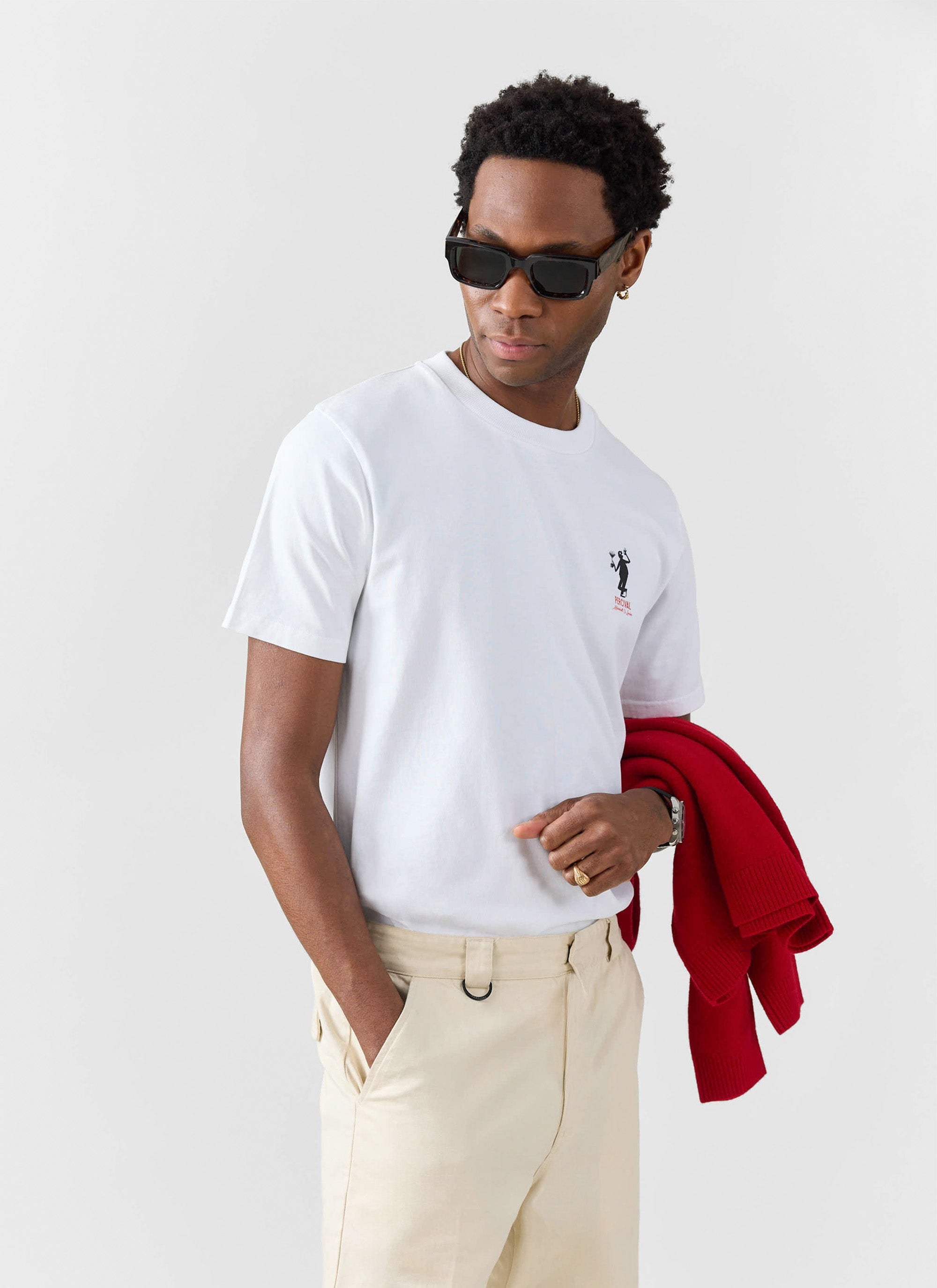 A model wearing sunglasses and the white Percival Carlo Mixologist t-shirt with beige trousers, holding a red jumper.