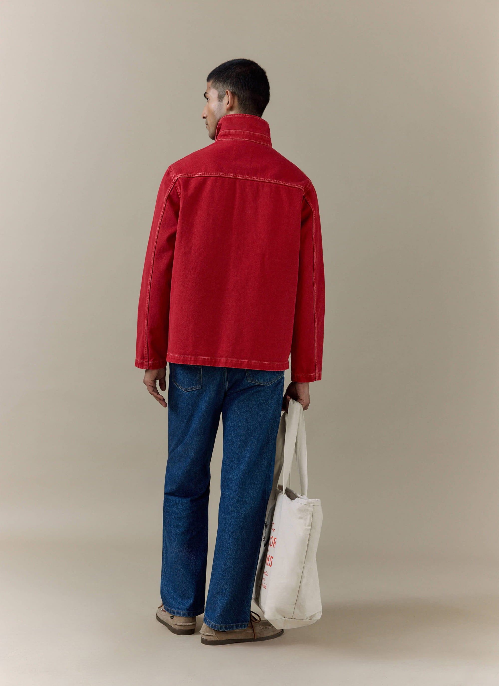 Rear view of the red cotton canvas Percival Oliver Chore Jacket, styled with blue jeans and a tote bag.