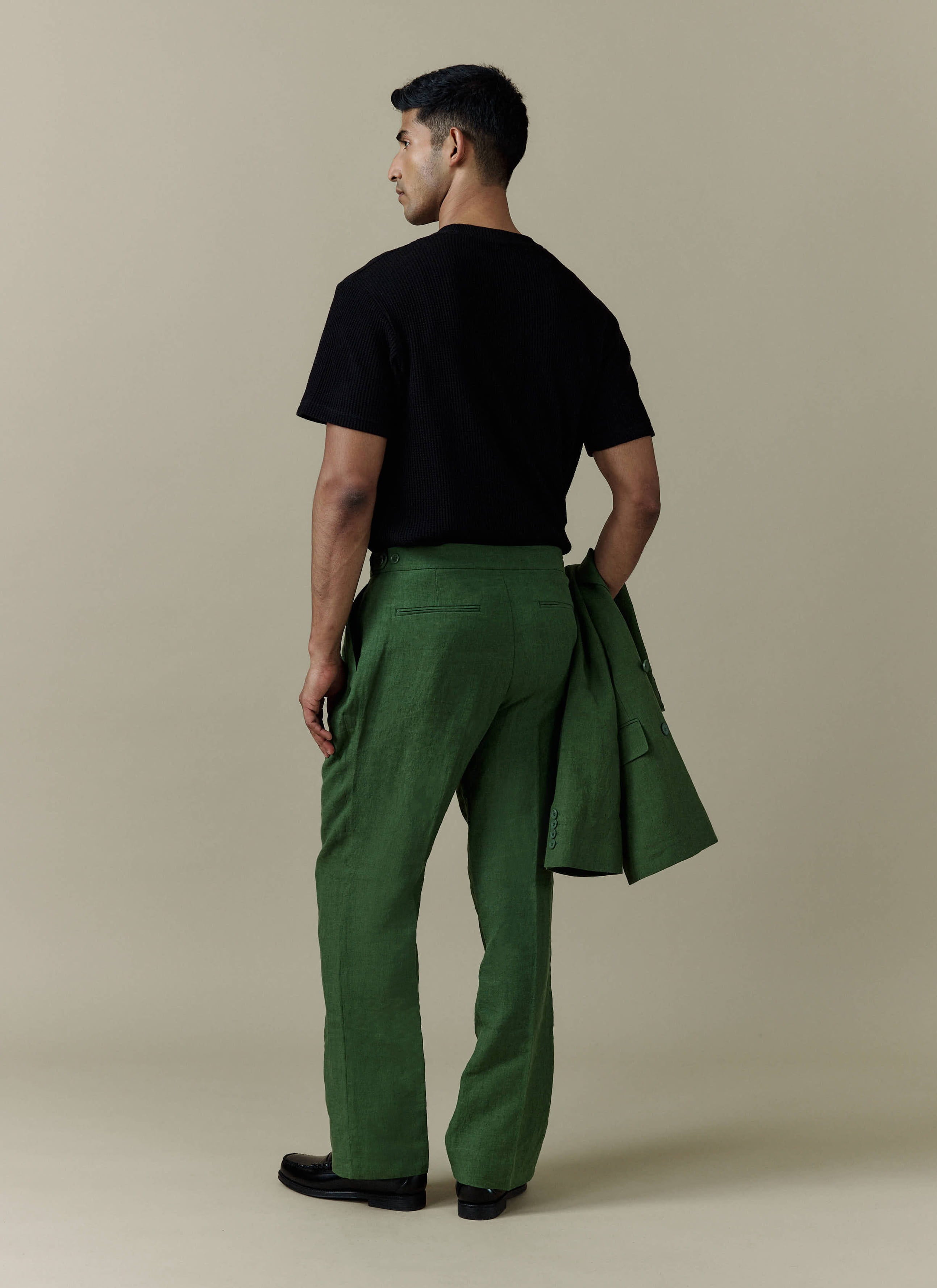 A model from behind in Percival forest green pleated linen trousers, holding a matching tailored linen blazer.