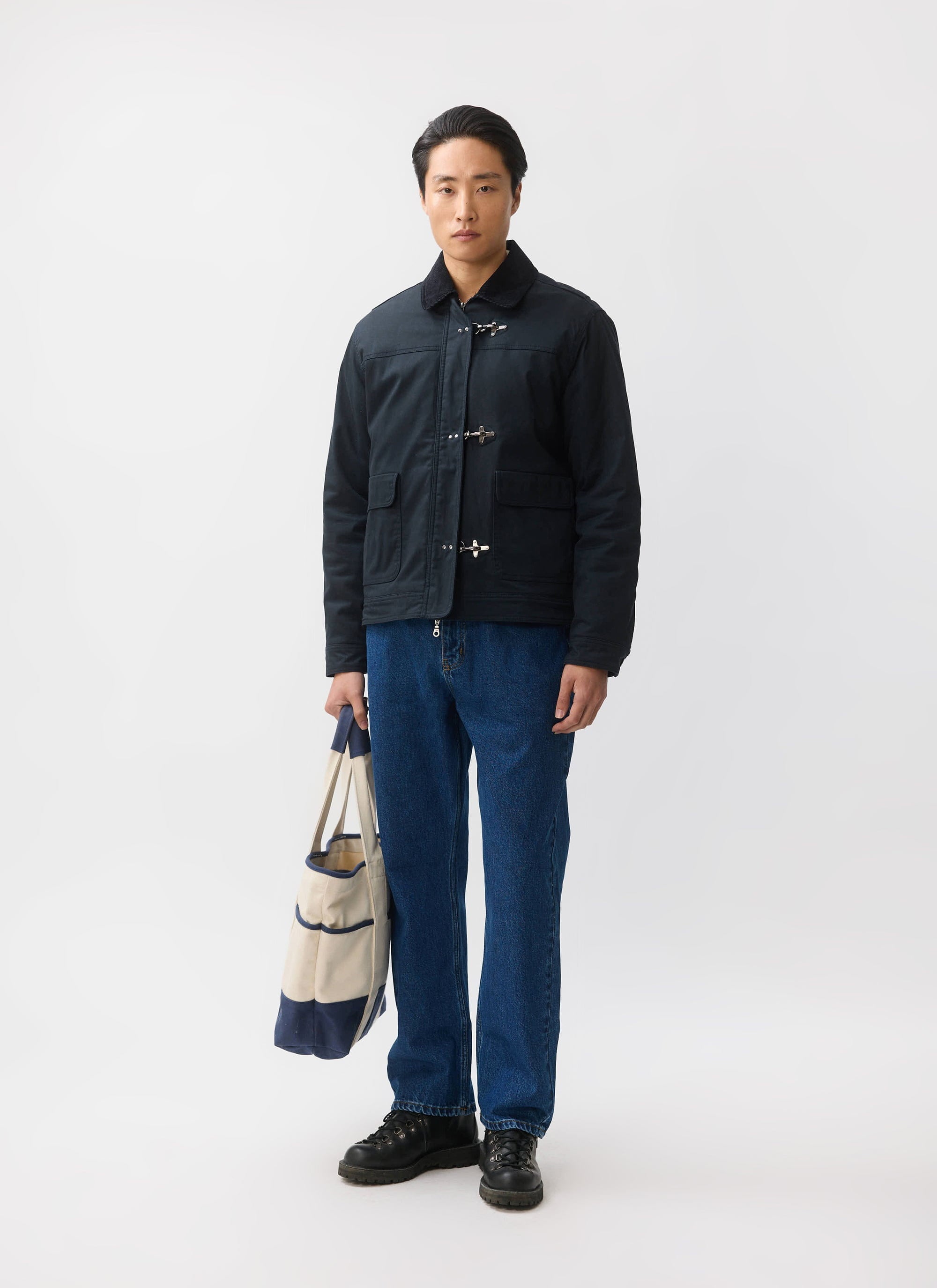 A model wears the navy Fontane Wax Jacket from Percival, paired with blue jeans and black boots, holding a tote bag.