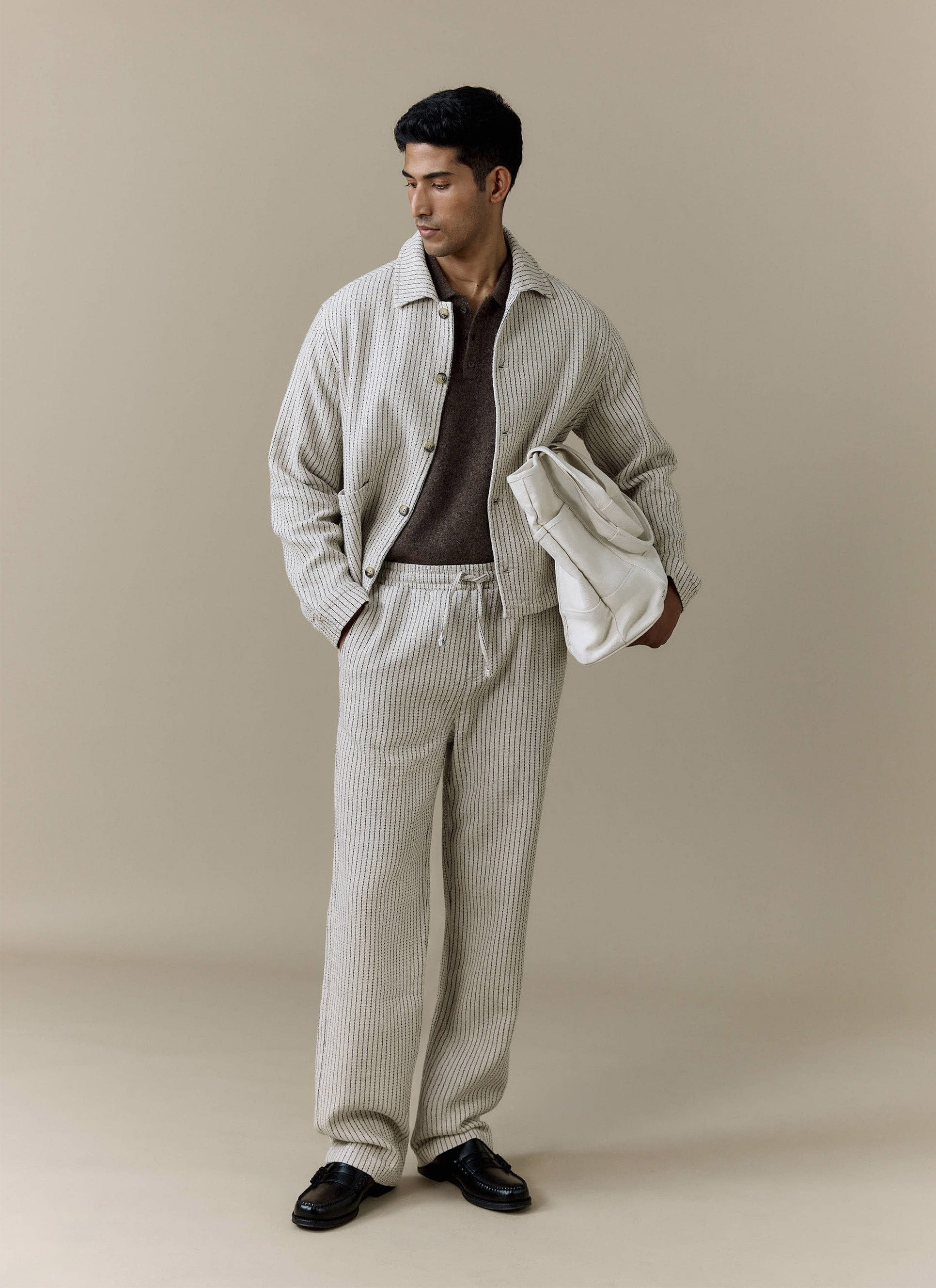 A model wears the Percival Sorrento Boxy Overshirt and matching trousers in ecru linen, styled with a brown polo shirt.