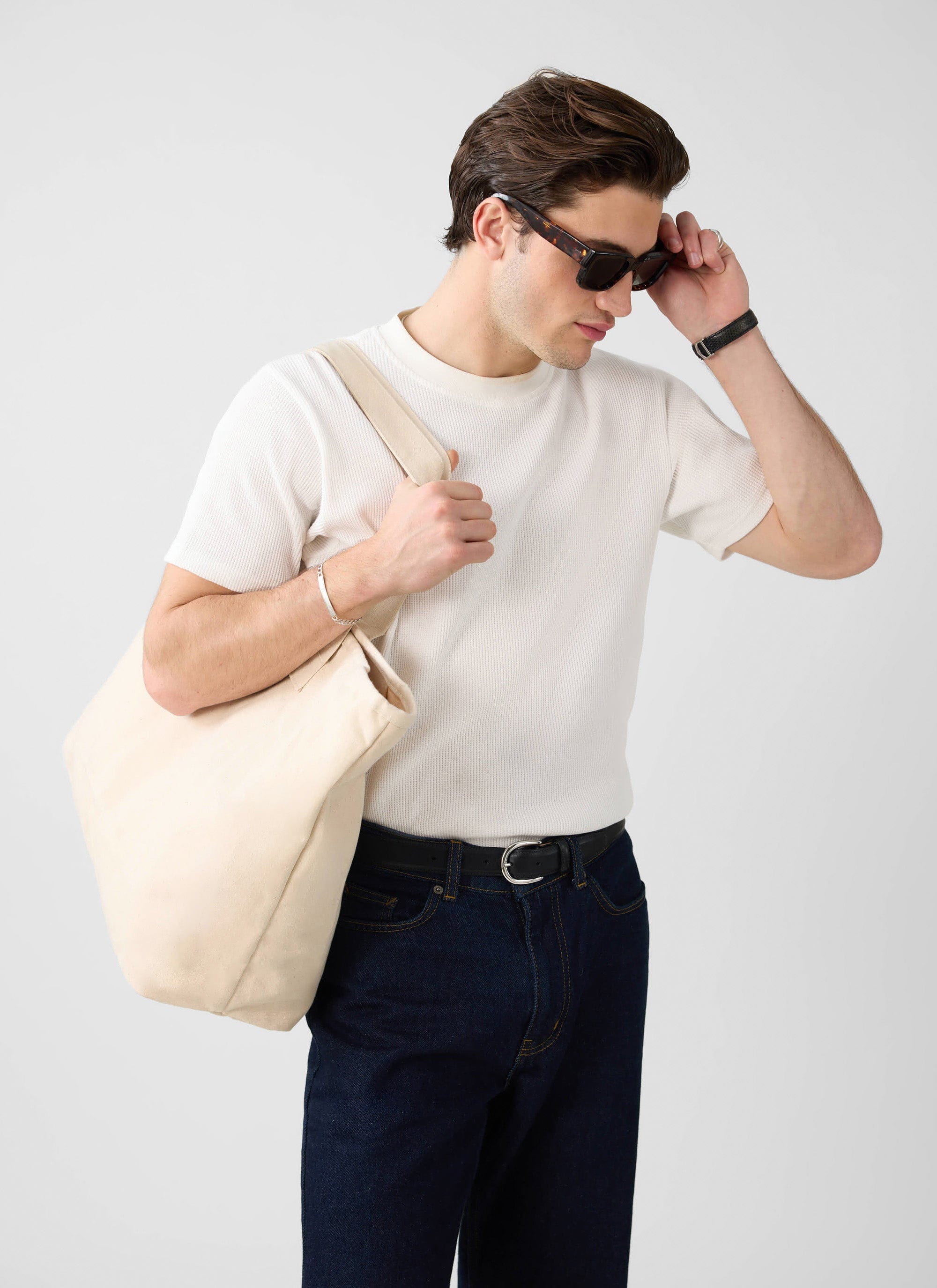 A man models the Percival white Waffle Short Sleeve T-Shirt, styled with dark jeans, a tote bag and sunglasses.