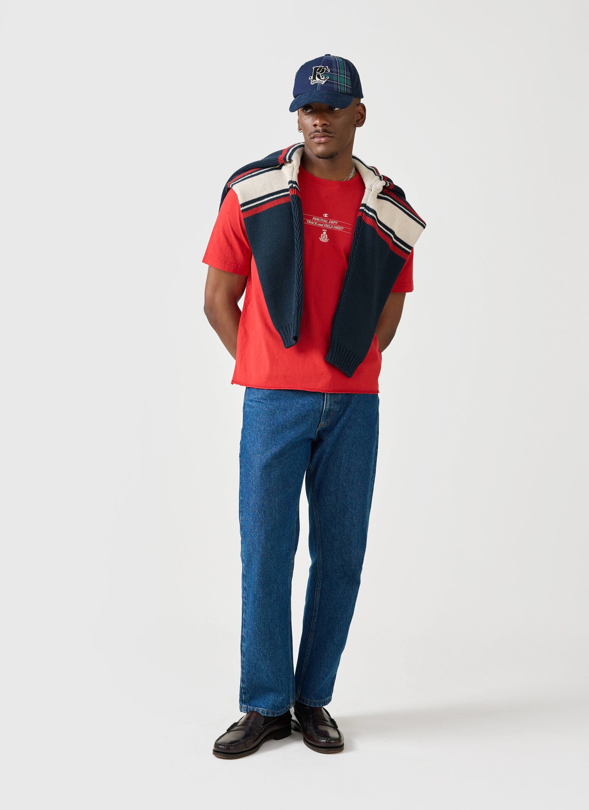 A model wears the red Track and Field T-Shirt from the Champion x Percival collection, styled with jeans and striped jumper.