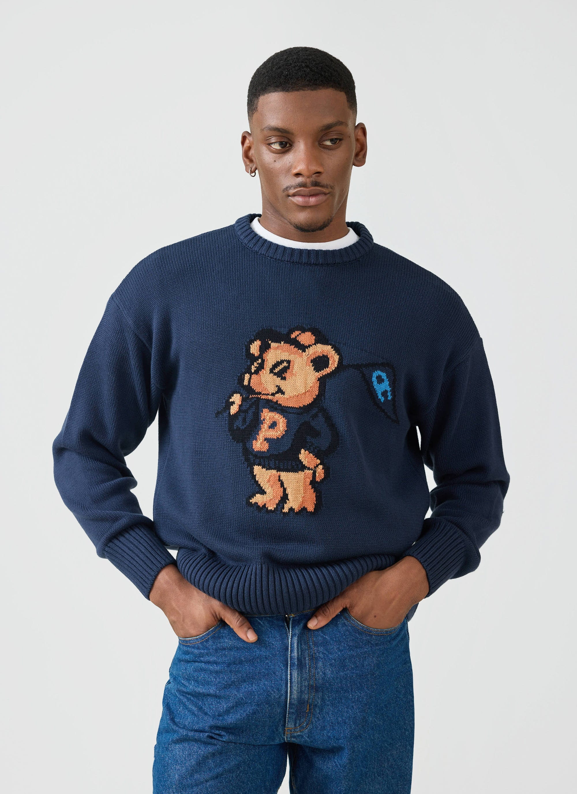 A man models the navy blue Jacquard Bear Crew Neck from Percival, a collaboration with Champion, styled with blue jeans.