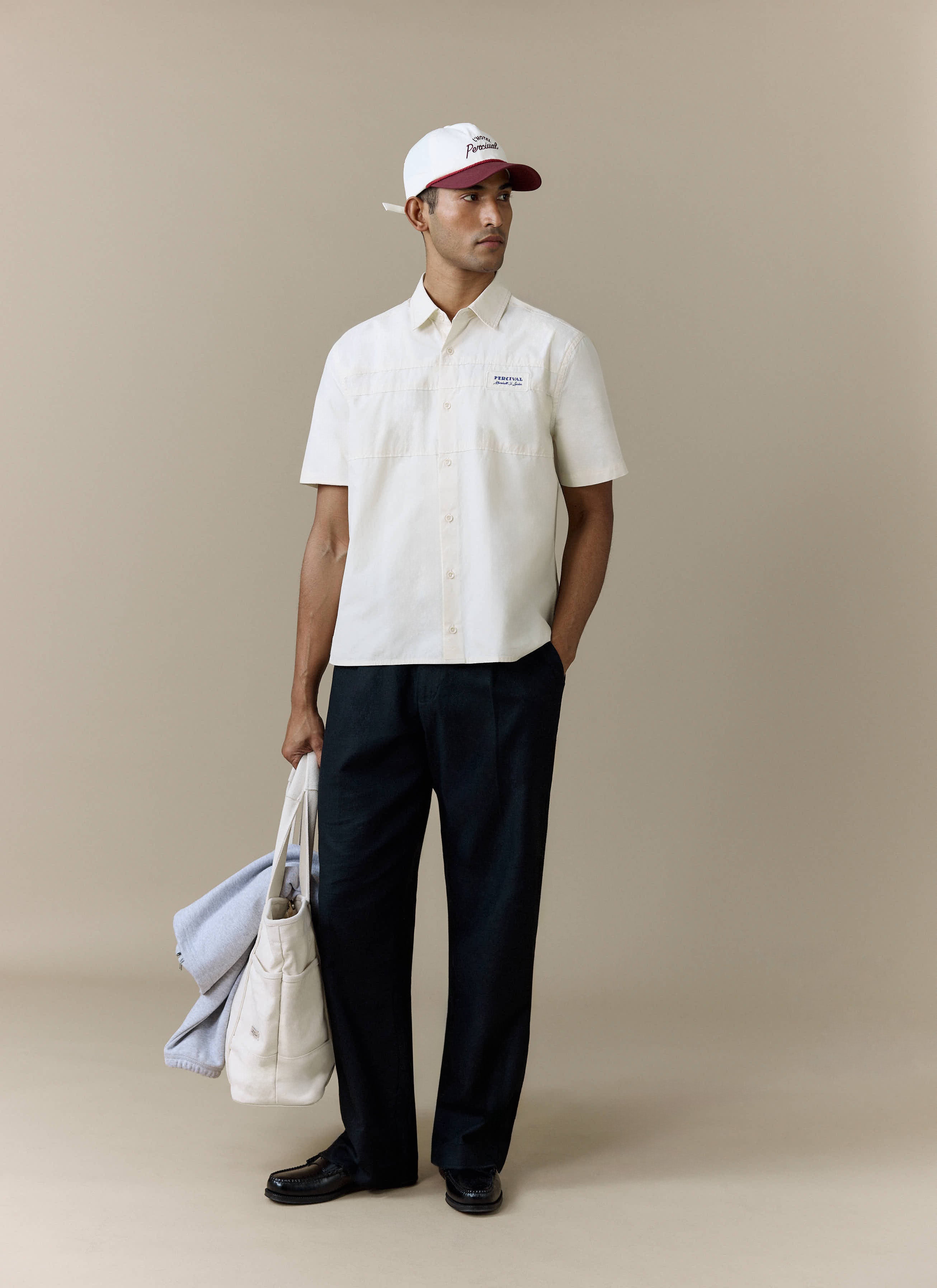 A full-length shot of a model wearing the Percival Harlow Boxy Shirt in ecru, styled with black trousers and a cap.